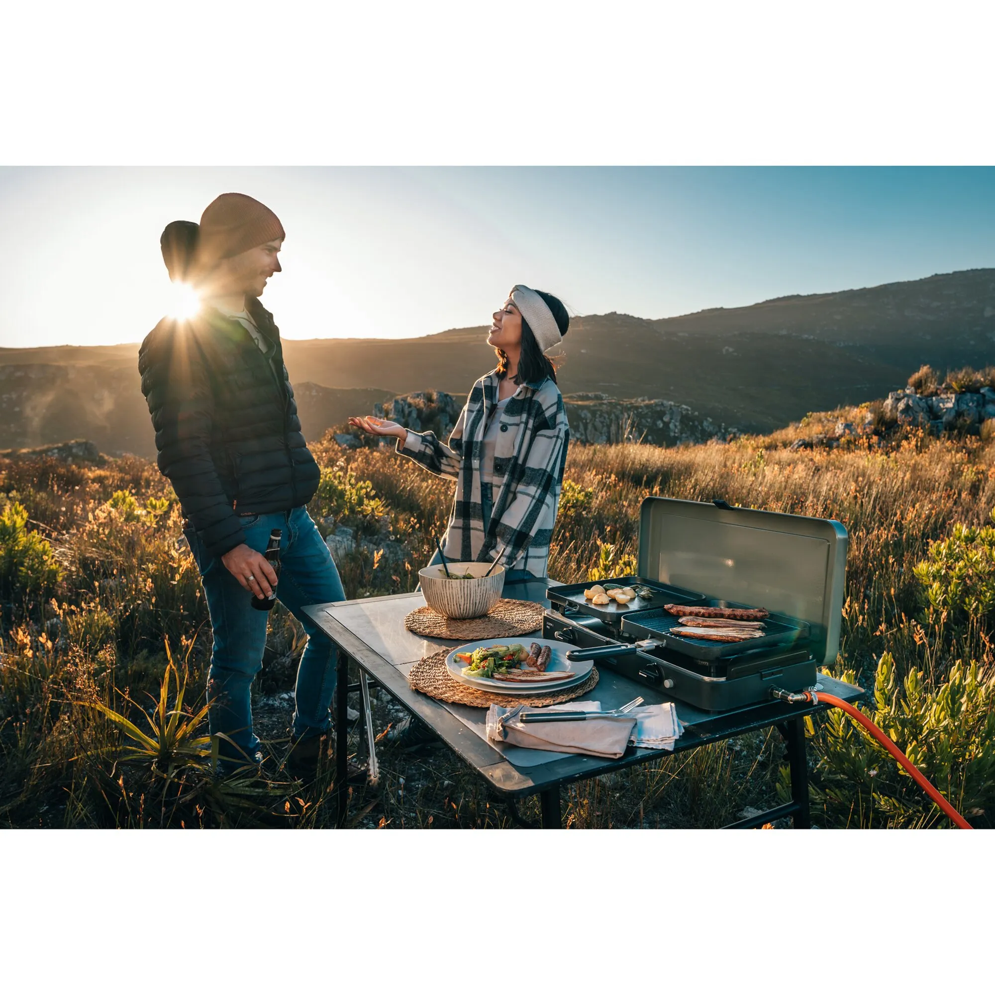 Zwei Menschen kochen in Berglandschaft am tragbaren Gaskocher 2 Cook 3 Pro Stove mit Grill und zubereiteten Speisen