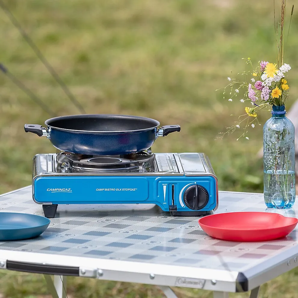 Blauer Campingaz Camp’Bistro DLX Stopgaz mit schwarzer Pfanne auf einem Campingtisch im Freien neben Geschirr und Blumenvasen
