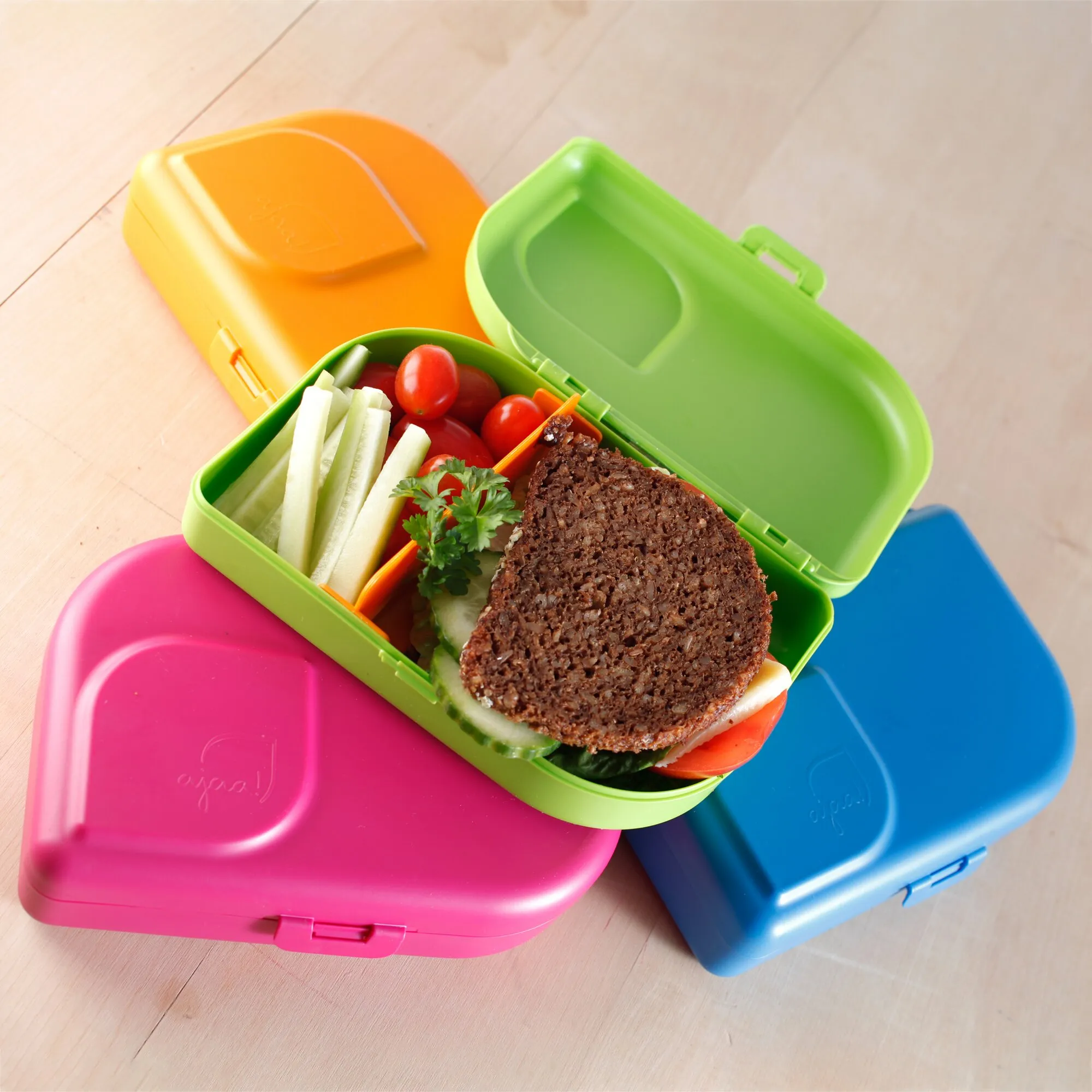 Blaue Brotbox neben pinken und orangefarbenen Boxen, geöffnet mit Vollkornbrot, Gurken, Tomaten und Gemüse gefüllt