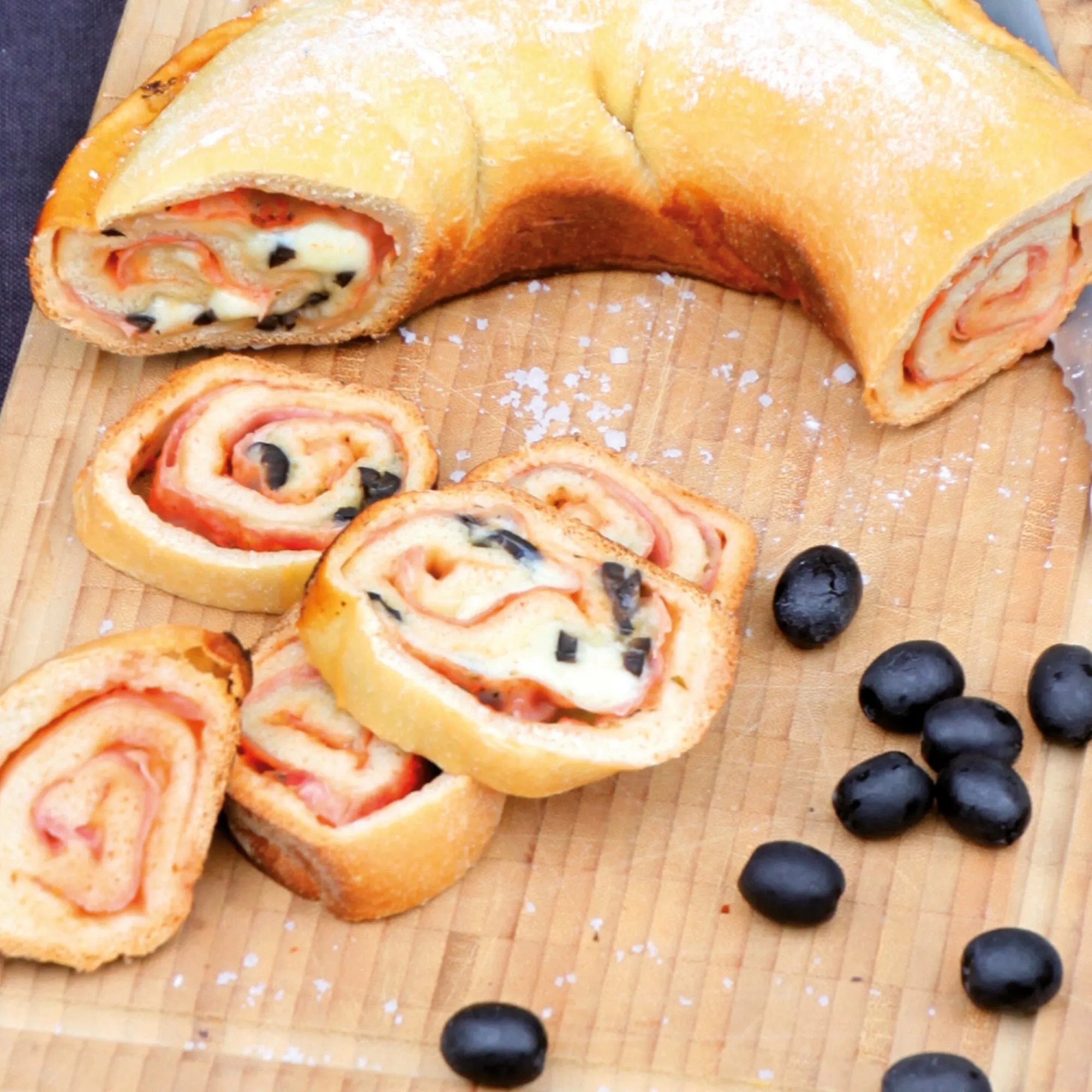 Herzhafte Blätterteig-Schnecken mit Schinken, Käse, Tomaten und schwarzen Oliven auf einem Holzbrett aus dem Omnia-Kochbuch