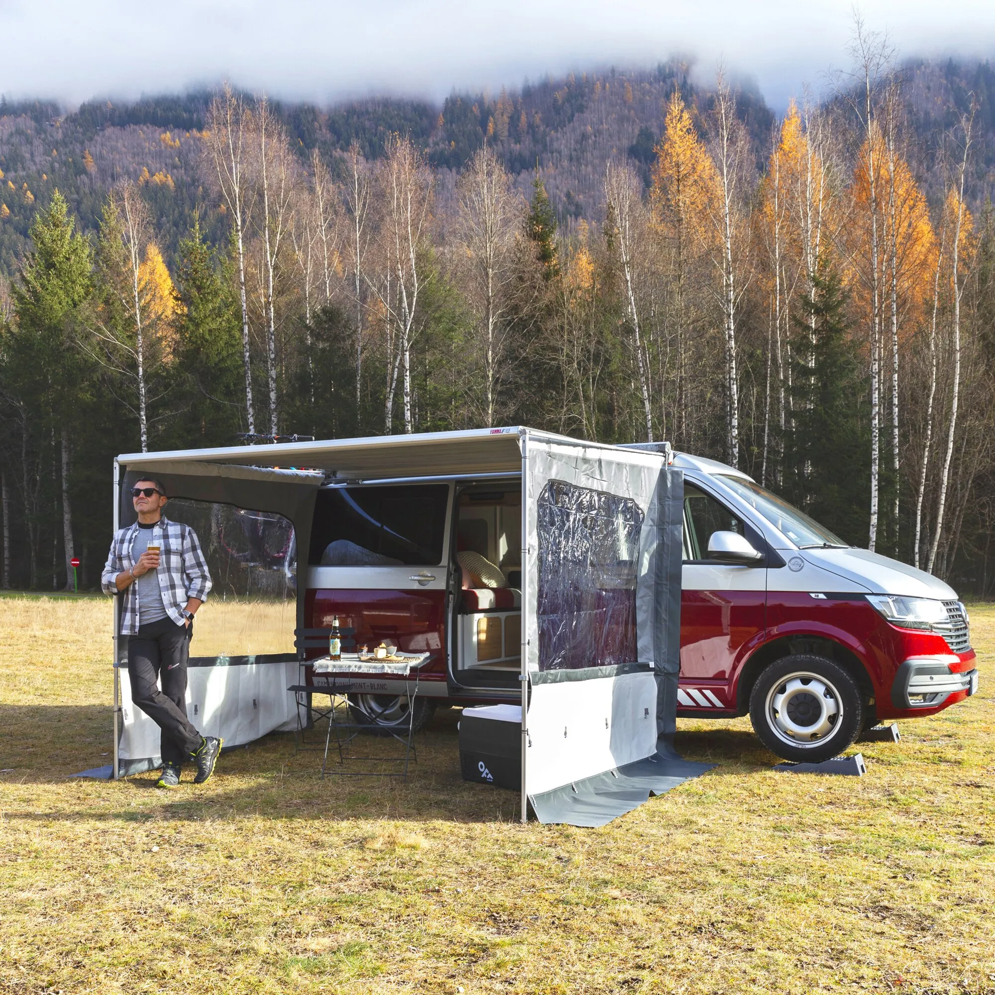 Roter Campervan mit ausgefahrener F35-Markise und geschlossener Seitenwand Side W Pro, davor ein Mann im Herbstwald gras.