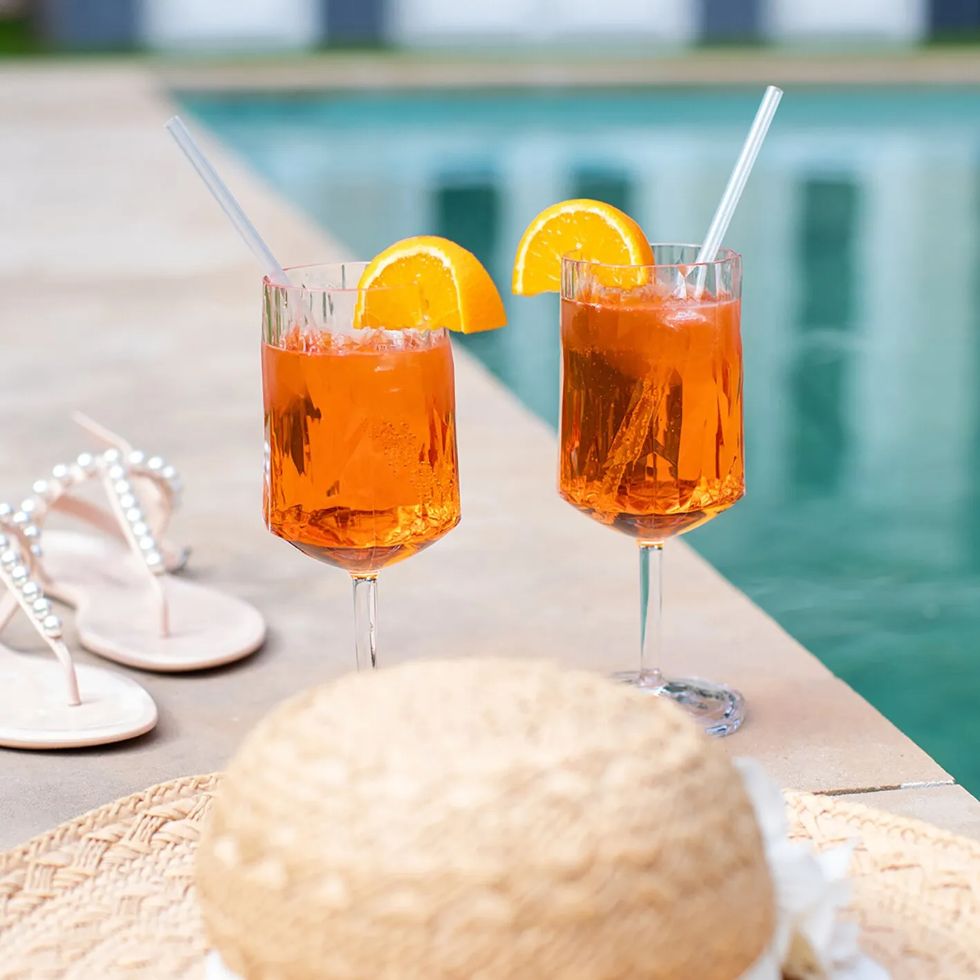 Zwei transparente Rotweingläser Superglas CLUB mit orangefarbenem Eisgetränk, Orangenscheiben und Trinkhalmen am Pool