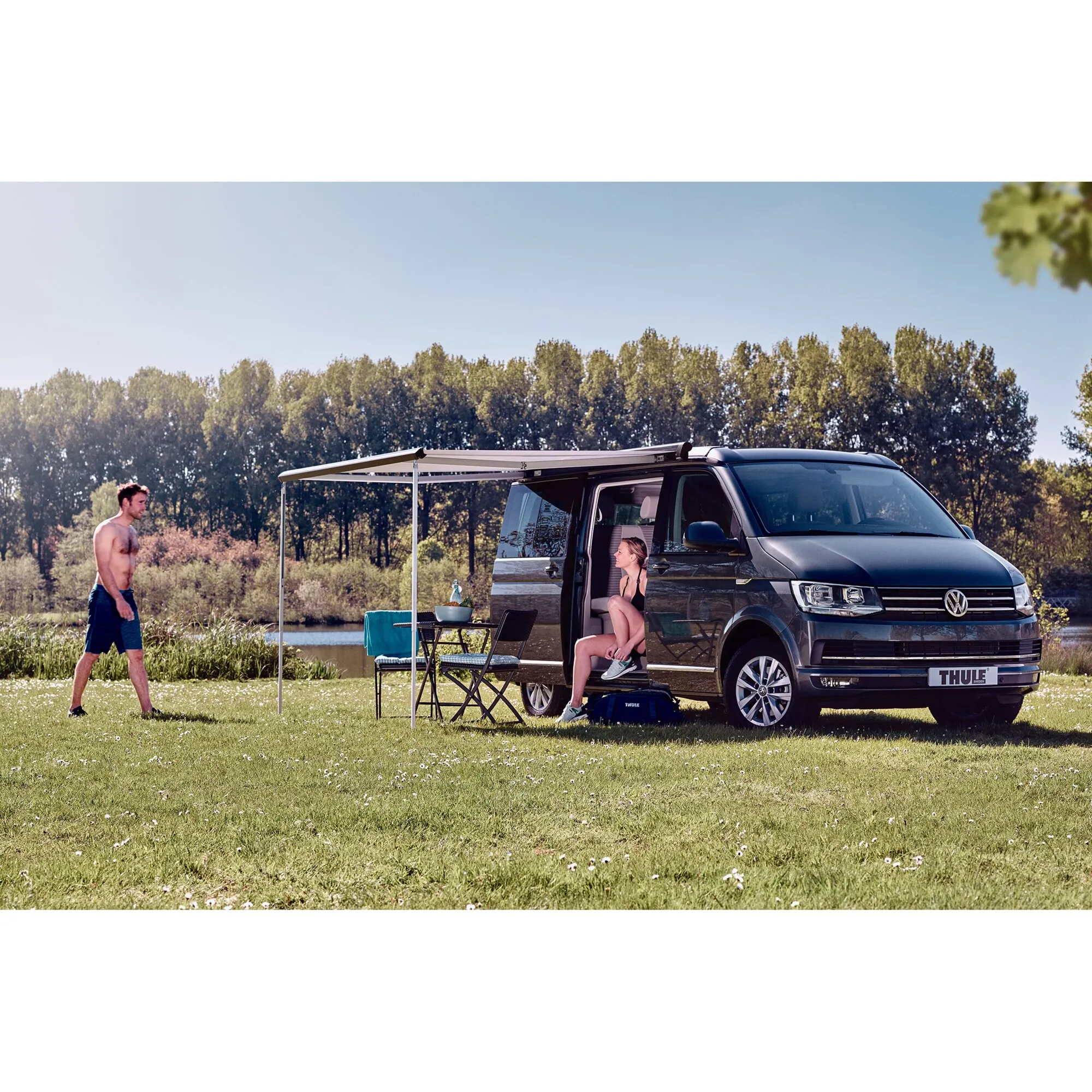 Hellgraue Thule Omnistor 5102 Markise an einem schwarzen Campervan, ausgefahren mit Stützstangen, Tisch und Stühlen am See