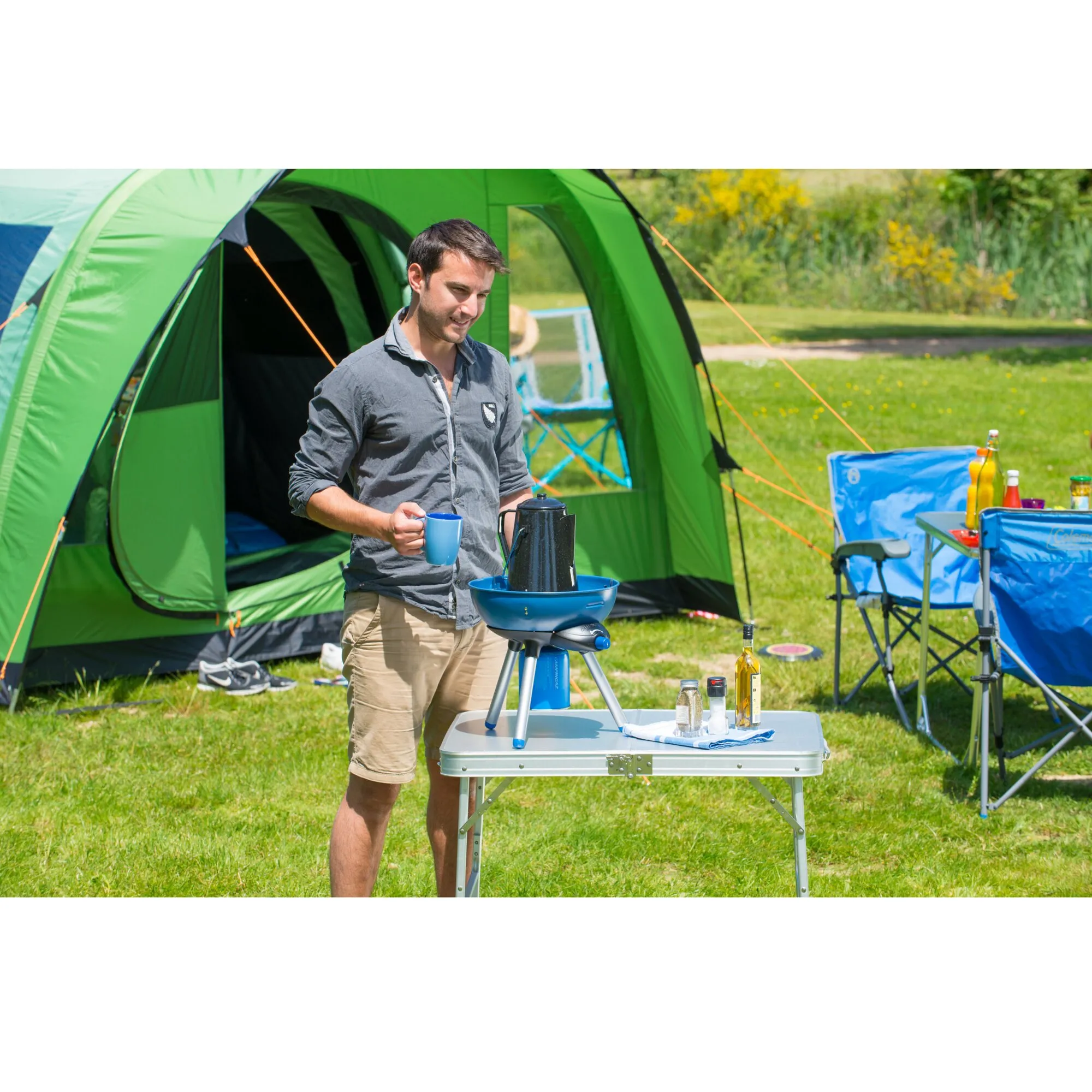 Blauer Party Grill 200 CV auf Dreibein beim Camping vor einem grünen Zelt, daneben ein Mann mit Tasse