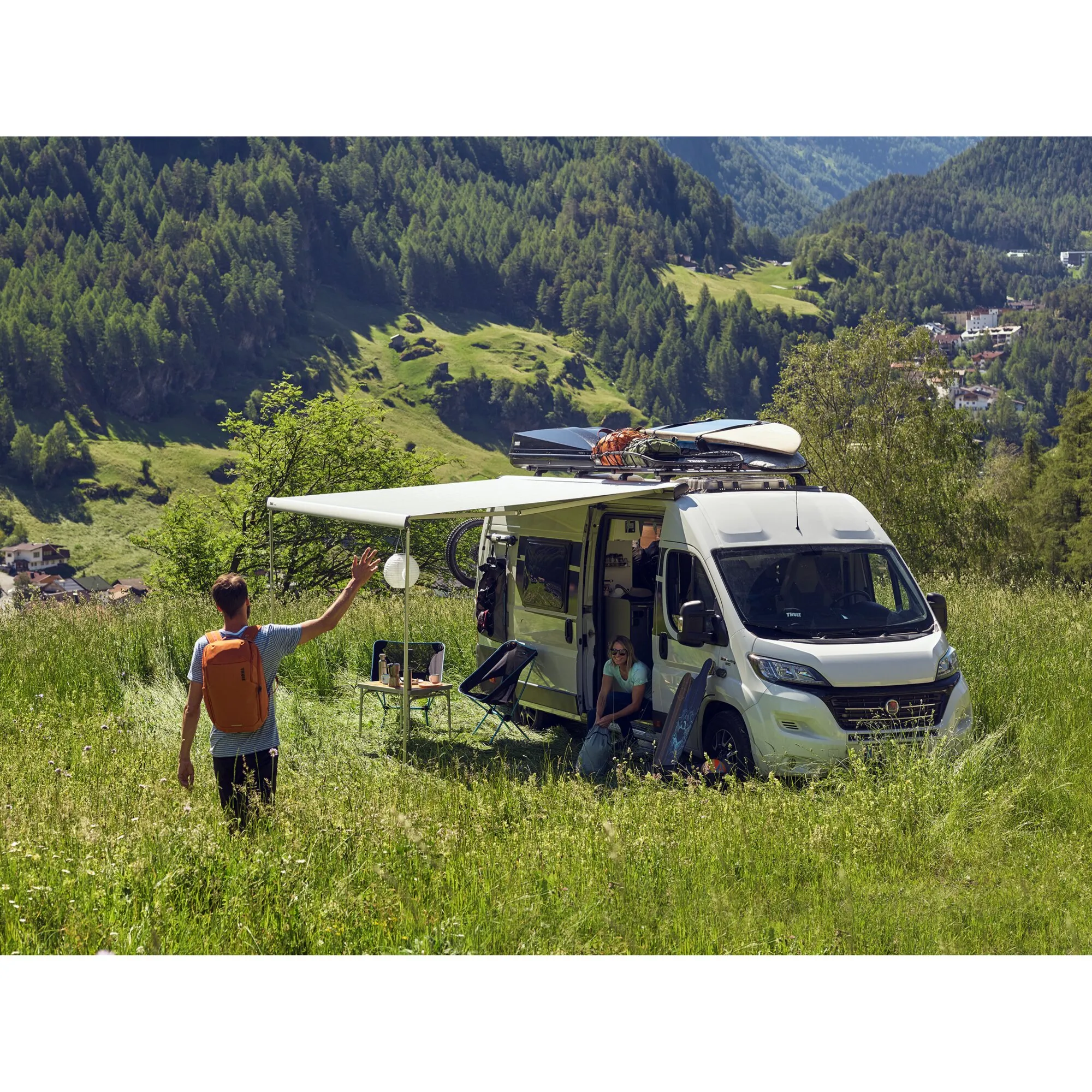 Weißer Fiat Ducato mit ausgefahrener grauer Thule Omnistor 6300 Markise auf einer Bergwiese, zwei Personen und Campingausrüstung
