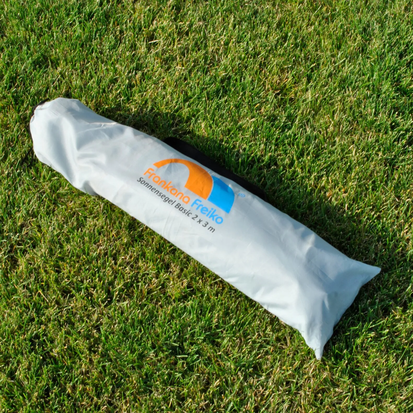 Weiße Tragetasche mit orange-blauem Frankana Freiko Logo und Aufdruck Sonnensegel Basic 2 x 3 m auf einer Rasenfläche