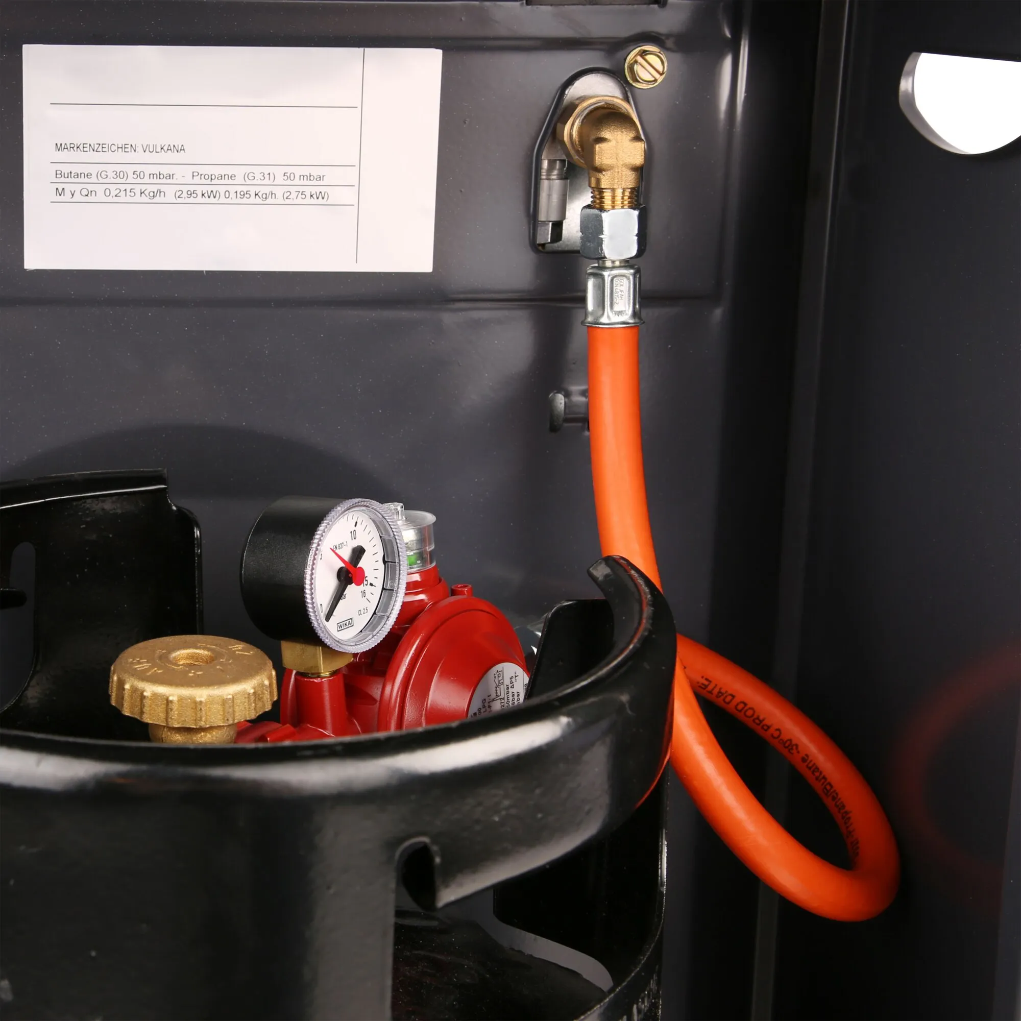 Detailansicht eines In-/Outdoor-Anschlusssets mit orangefarbenem Gasschlauch, Messinganschluss und rotem Druckregler mit Manometer in einem schwarzen Gehäuse