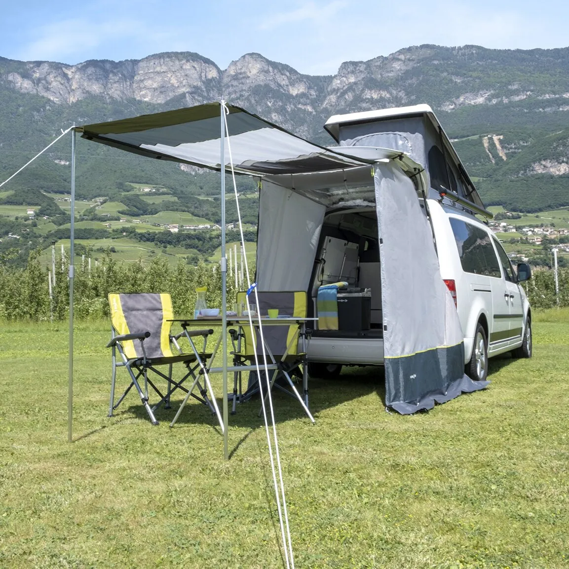 Heckzelt Pilote Caddy 4 an weißem Campingbus mit Sonnendach, zwei Campingstühlen und Tisch vor einer Berglandschaft