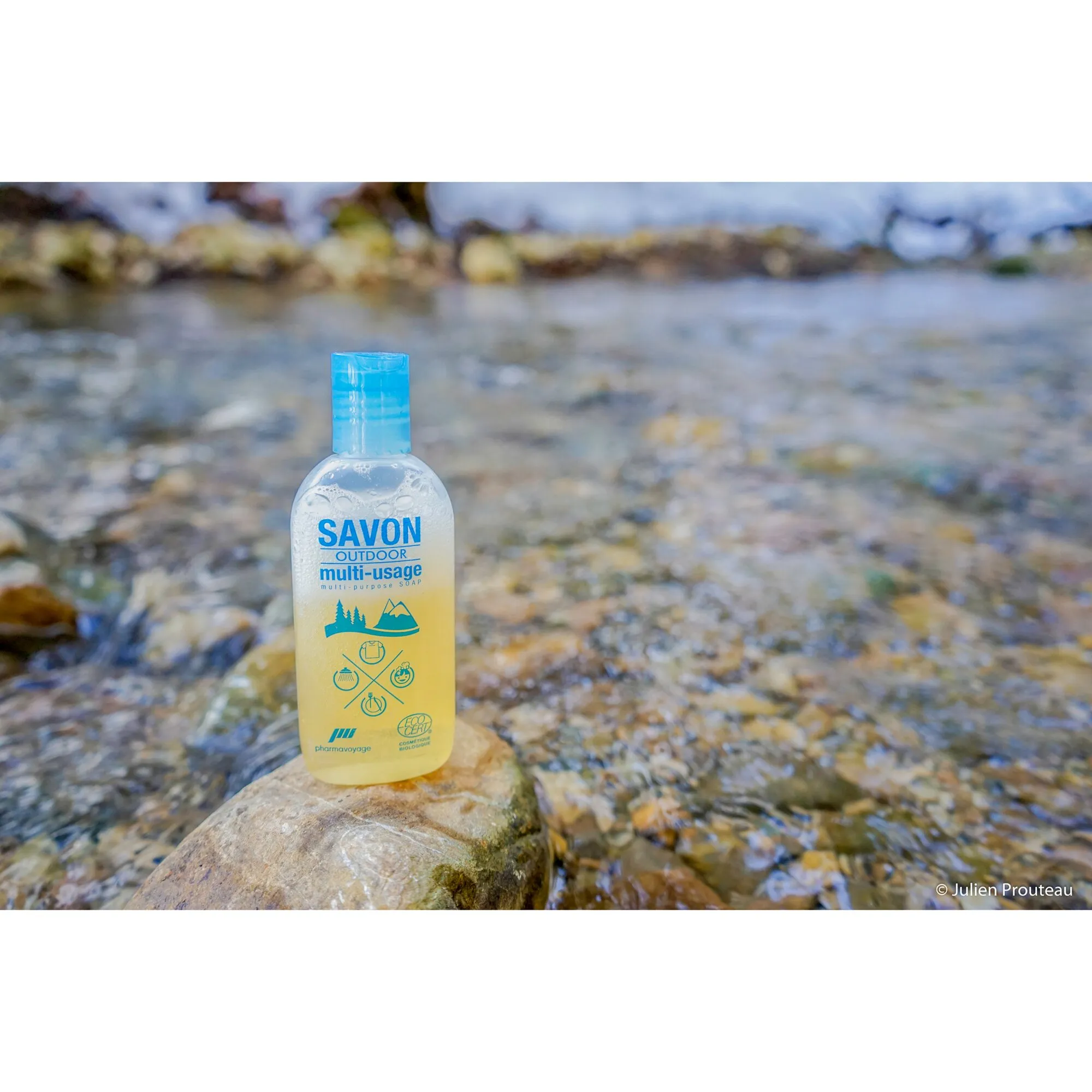 Transparente Flasche mit blauer Kappe und gelber Flüssigseife SAVON OUTDOOR multi-usage auf einem Stein in einem Bachbett