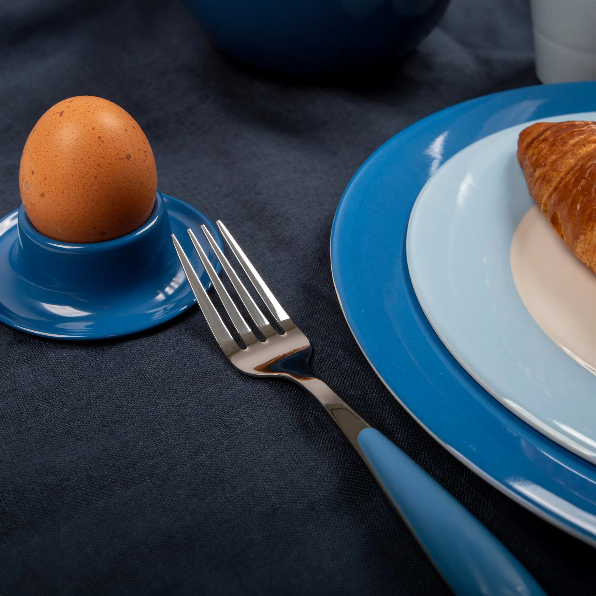 Blaues Geschirrset Colour Line mit Teller, Eierbecher und Tasse, angerichtet mit Ei und Croissant sowie Gabel auf dunkler Tischdecke