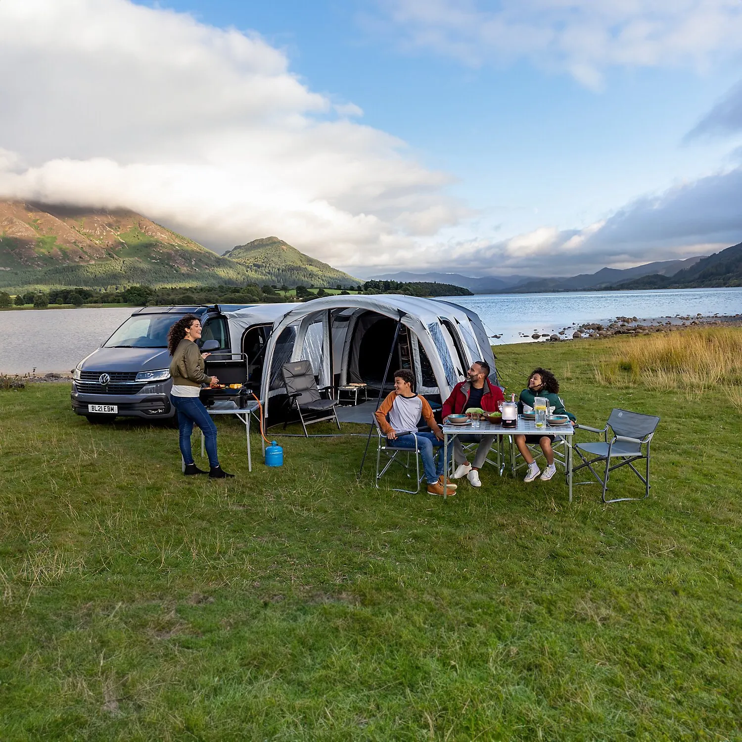 Graues Busvorzelt Journeymaster Deluxe L an einem VW-Camper mit Sitzbereich, Tisch und vier Personen beim Essen am See
