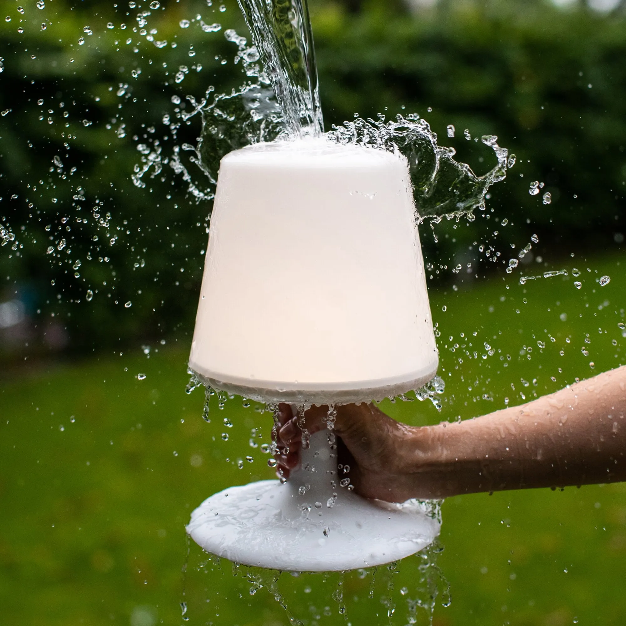 Weiße mobile Tischleuchte mit rundem Fuß und kegelförmigem Leuchtkörper, von Wasser bespritzt im Außenbereich