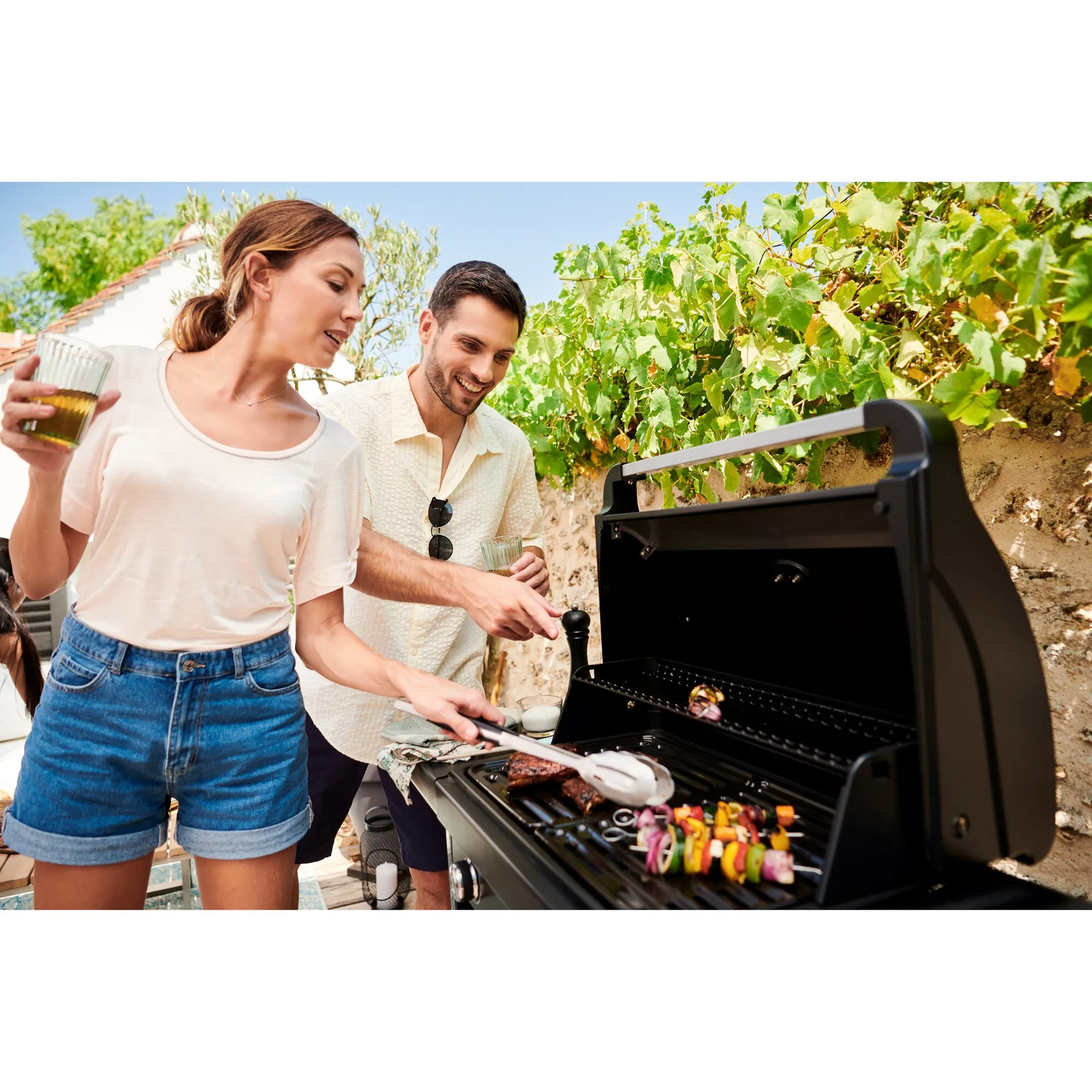 Offener schwarzer Gasgrill Compact 3 L mit Grillgut, während zwei Personen im Freien Gemüse und Fleisch grillen