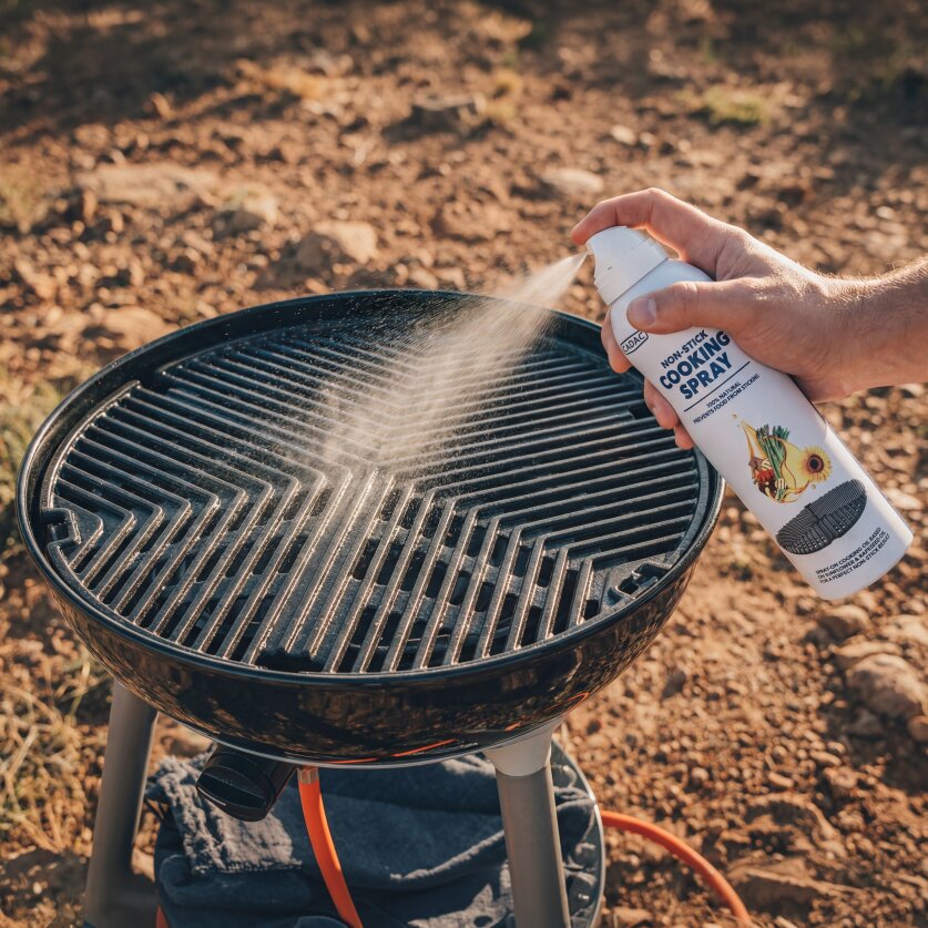 Eine Hand sprüht Antihaft-Kochspray aus einer weißen Dose auf die Grillroste eines runden Grills im Freien, Sprühnebel steigt sichtbar