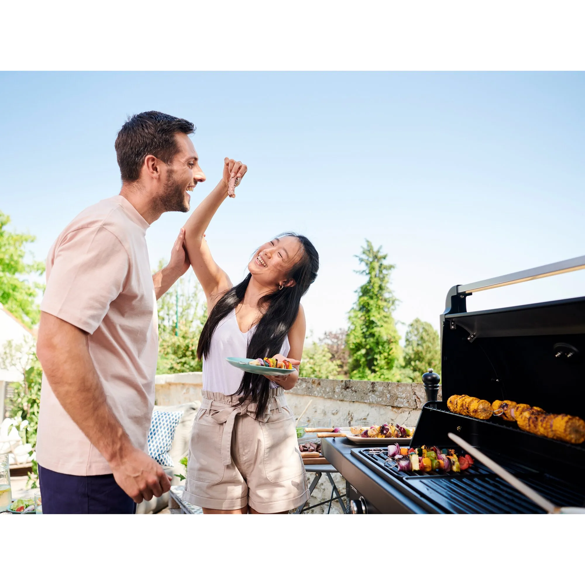 Paar beim Grillen im Freien neben geöffnetem schwarzem Gasgrill Compact 3 L mit Maiskolben und Gemüsespießen