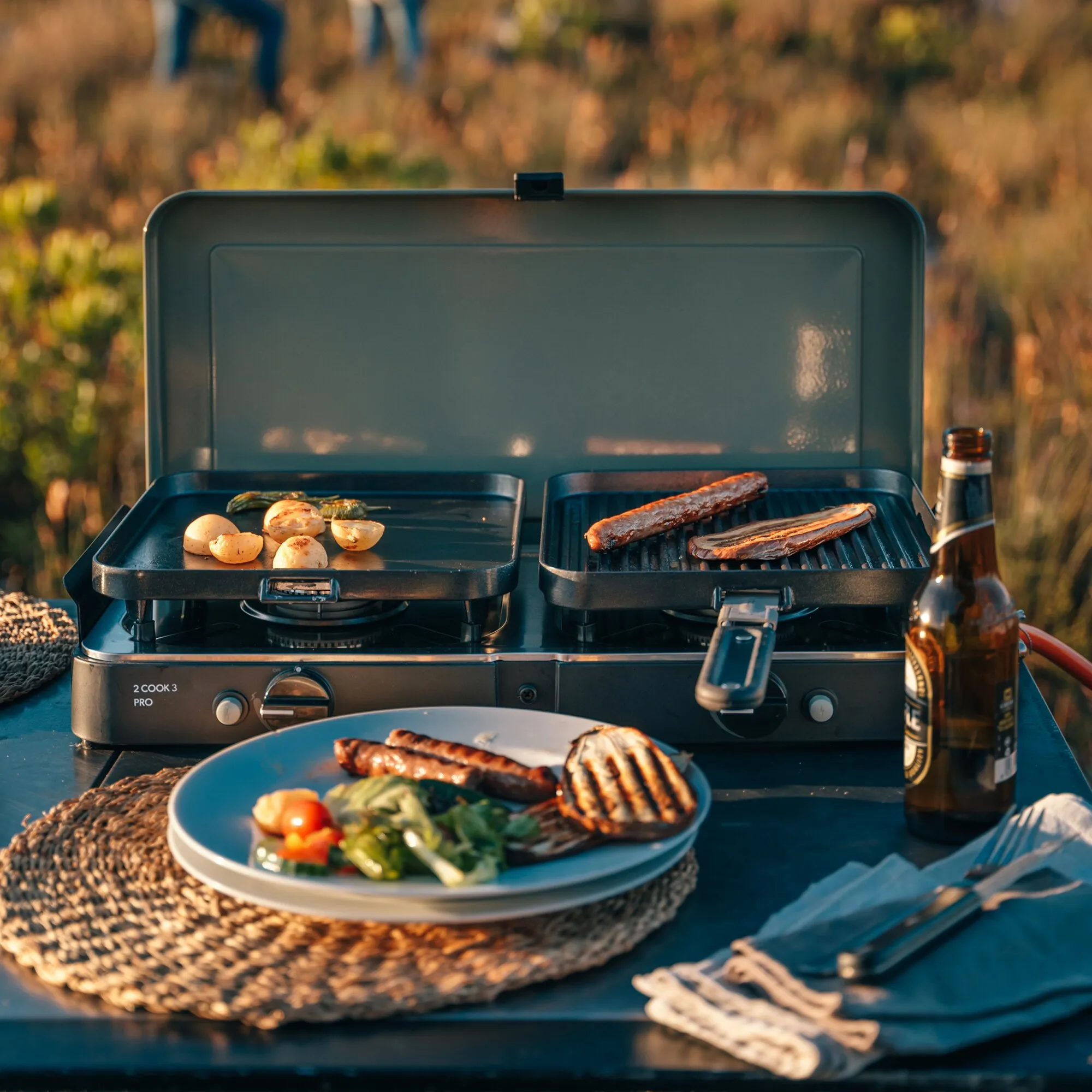 Gaskocher 2 Cook 3 Pro Stove mit zwei offenen Grillplatten, Würstchen und Gemüse beim Grillen im Freien