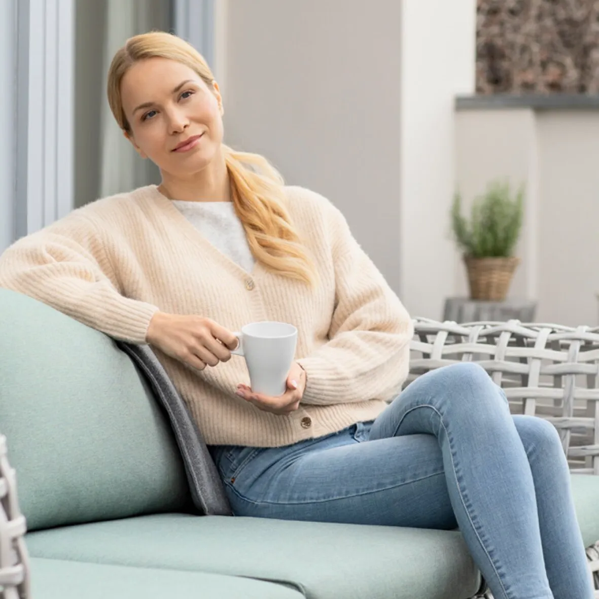 Frau sitzt mit einer Tasse auf einem Outdoor-Sofa und lehnt an einem grauen Heizkissen OL 750