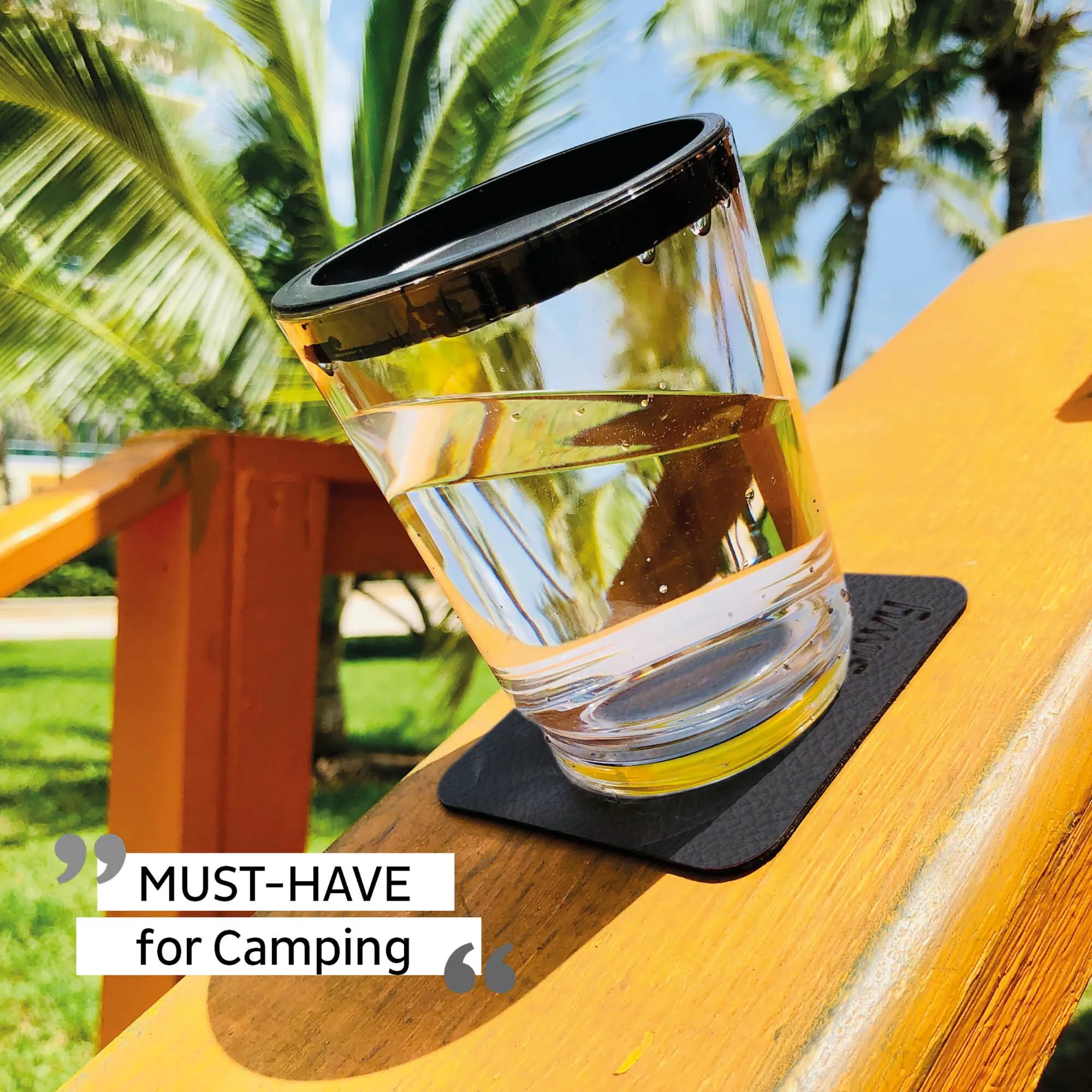 Transparenter Magnet-Trinkbecher mit schwarzem Rand auf einer Outdoor-Tischunterlage vor Palmen, Text: Must-Have for Camping