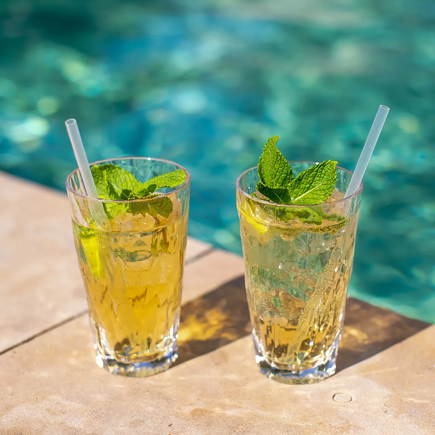 Zwei transparente Trinkgläser Superglas CLUB mit gelben Eisgetränken, Minzblättern und weißen Trinkhalmen am Pool
