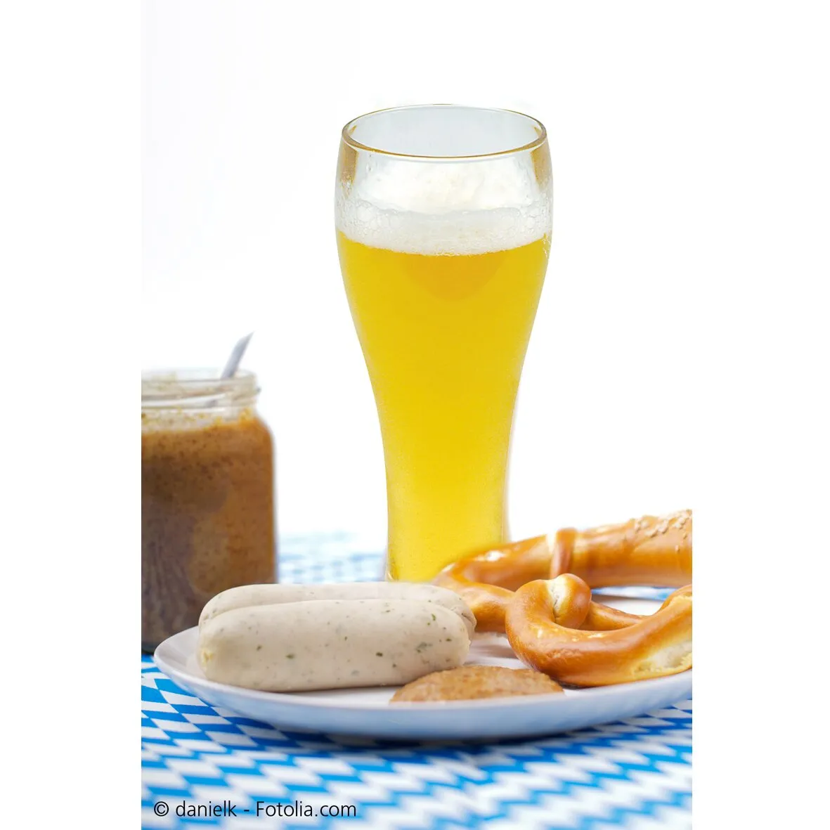 Weißbierglas aus klarem Glas, gefüllt mit goldenem Weißbier und weißer Schaumkrone neben Brezel und Weißwurst auf einem Teller