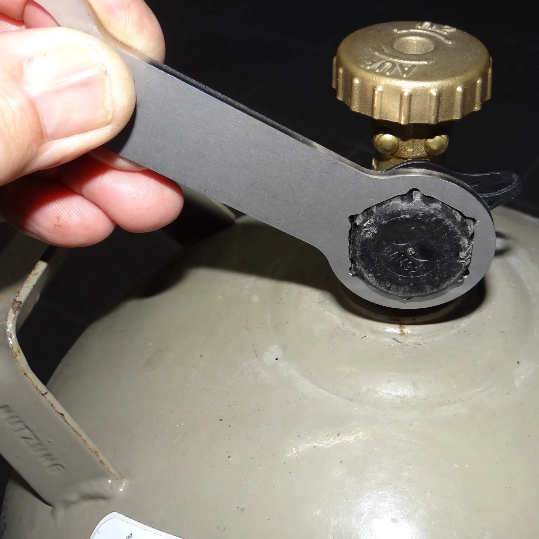 Gasflaschenschlüssel aus schwarzem Metall wird an einem Gasregler auf einer grauen Gasflasche angesetzt, daneben eine goldfarbene Ventilkappe