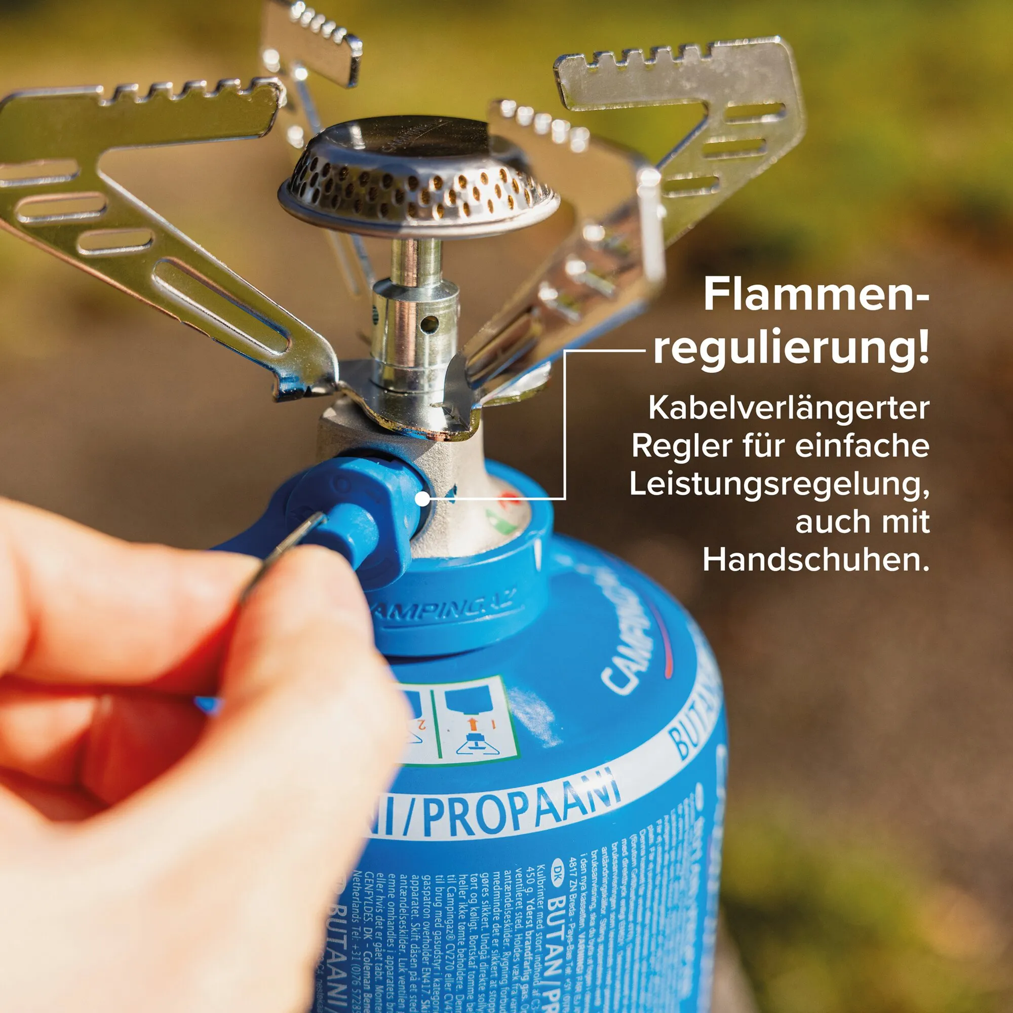 Detailaufnahme eines tragbaren Campingkochers mit perforiertem Brennerkopf, Metallträgern und blauer Propangasflasche; Hand greift nach dem Regler