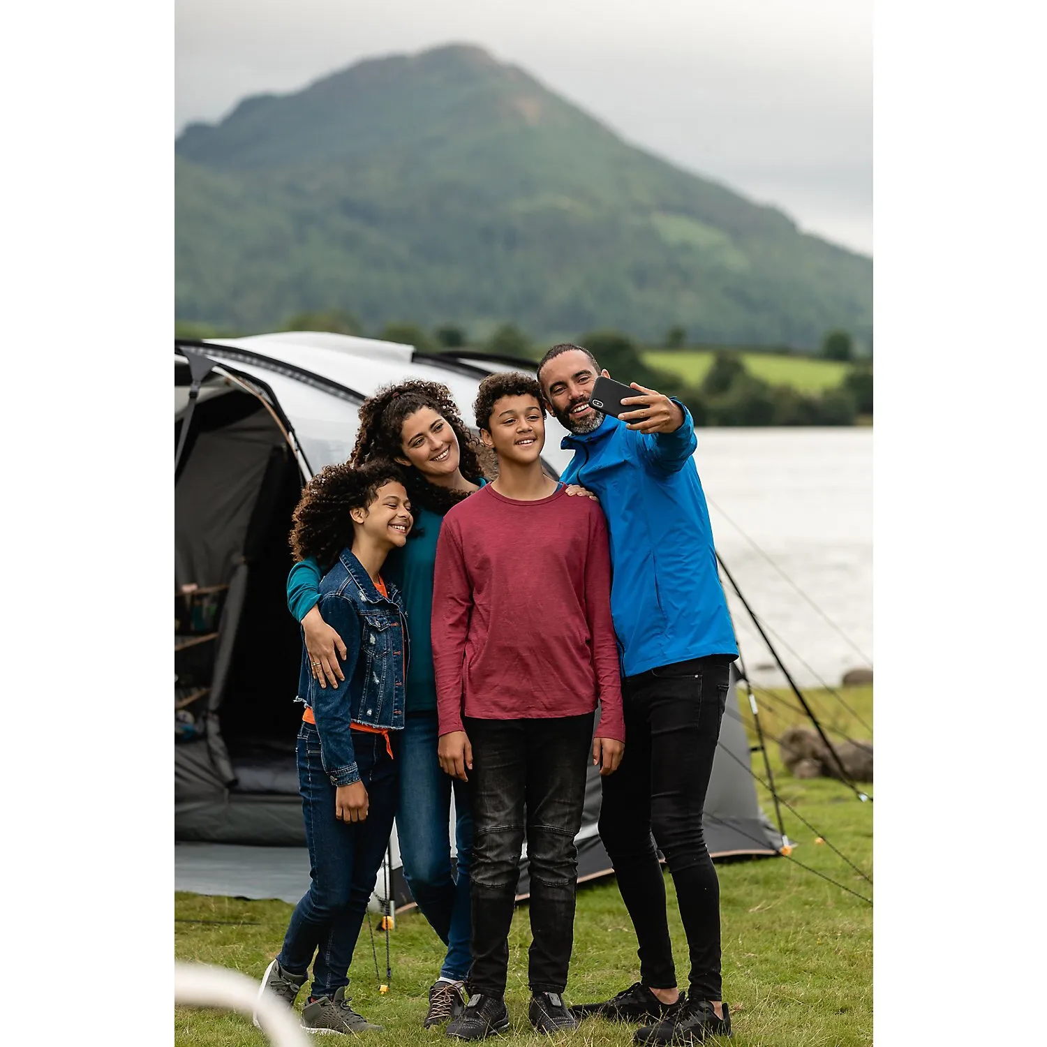 Familie macht vor dem Busvorzelt Journeymaster Deluxe L am See ein Selfie, mit Bergen und Landschaft im Hintergrund