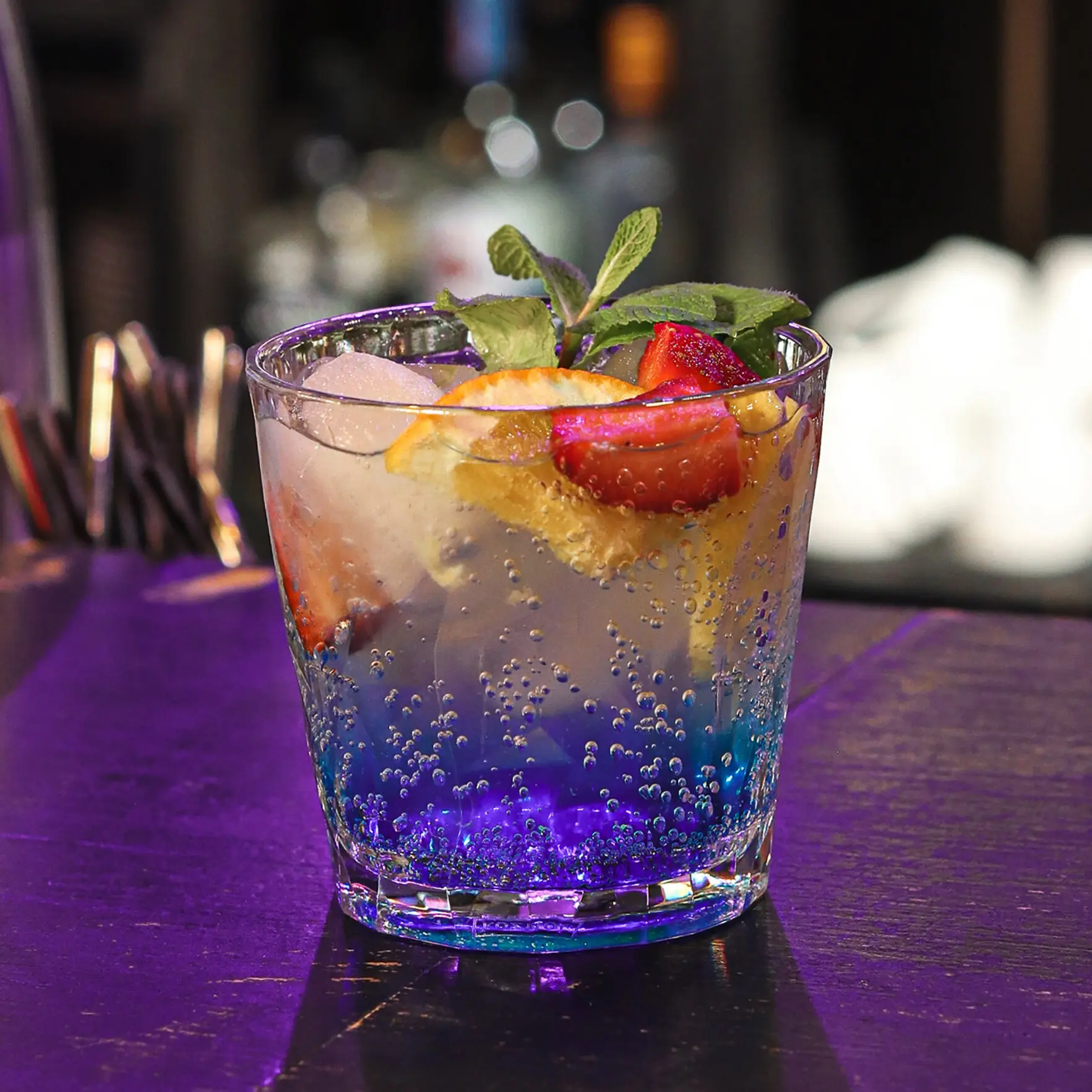 Trinkglas Superglas CLUB mit Sprudel, Eiswürfeln, Orangenscheibe, rote Obststücke und Minzblättern; blau-violetter Farbverlauf im Glas