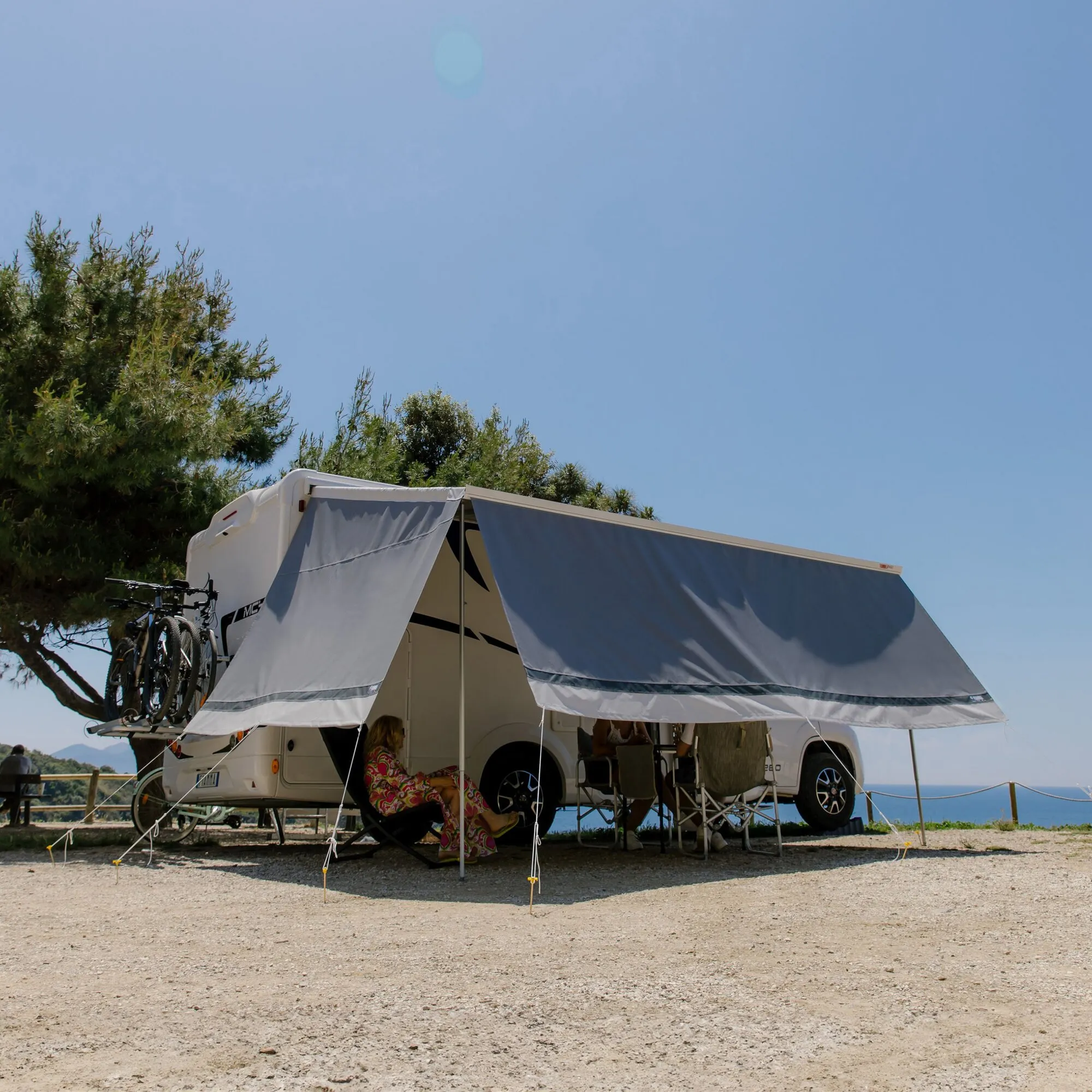 Wohnmobil mit ausgefahrener CaravanStore-Markise und grauen Seitenwänden auf einem Campingplatz am Meer, Fahrräder am Heck und Personen im Schattenbereich