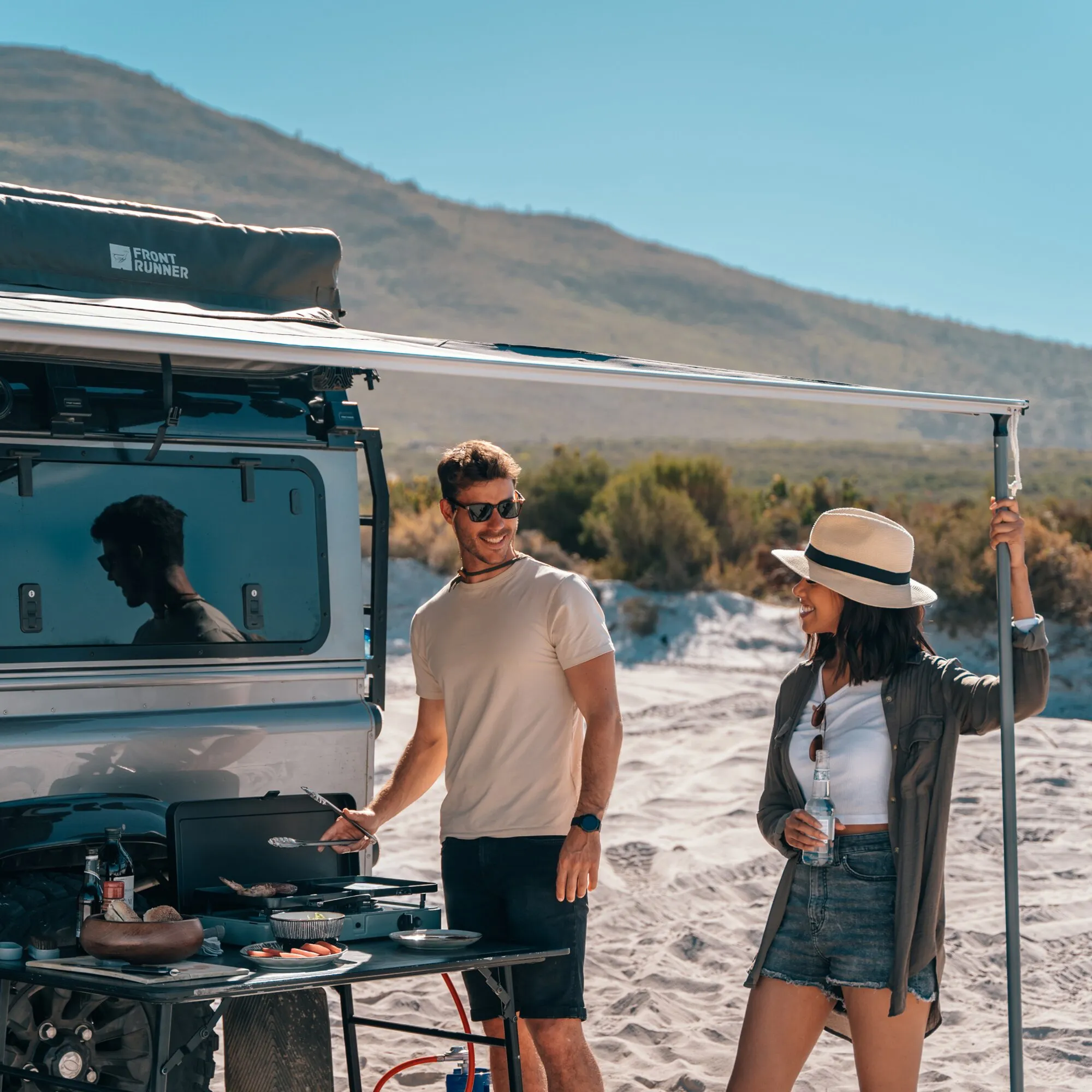 Gaskocher 2 Cook 3 Pro Stove beim Outdoor-Kochen am Fahrzeug mit Grillgut, Geschirr und zwei Personen