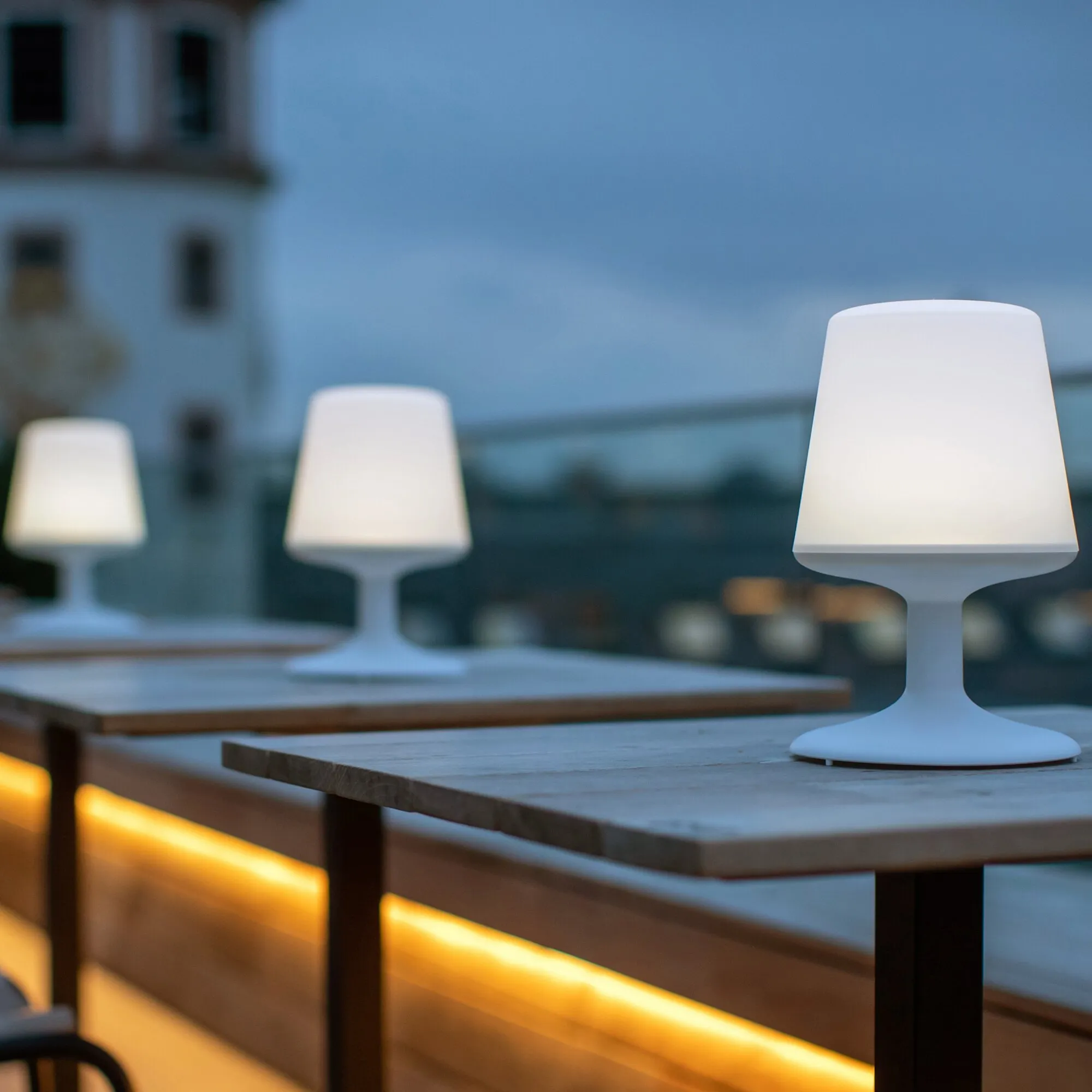 Weiße Tischleuchte Light to Go mit abgerundetem Schirm und breitem Standfuß auf einem Holztisch, zwei weitere Leuchten im Hintergrund