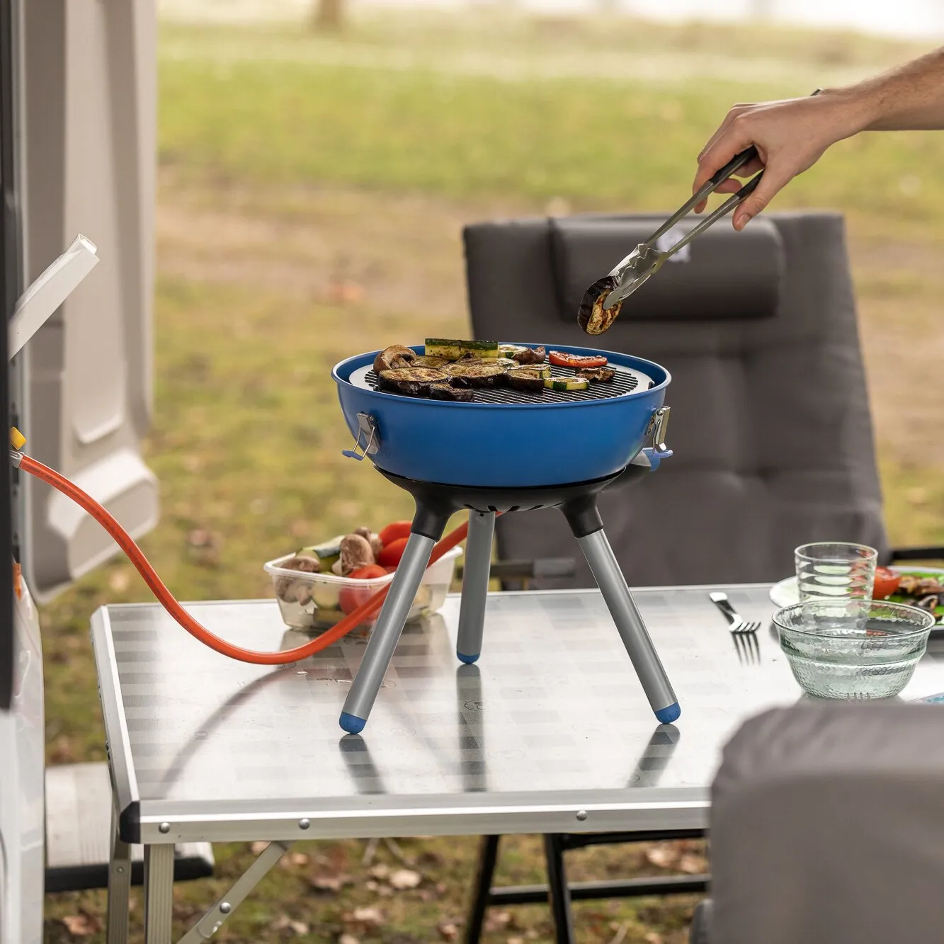 Blauer Party Grill 400 Caravan Connect mit Grillgemüse auf einem Campingtisch, während eine Hand Grillgut mit Zange wendet