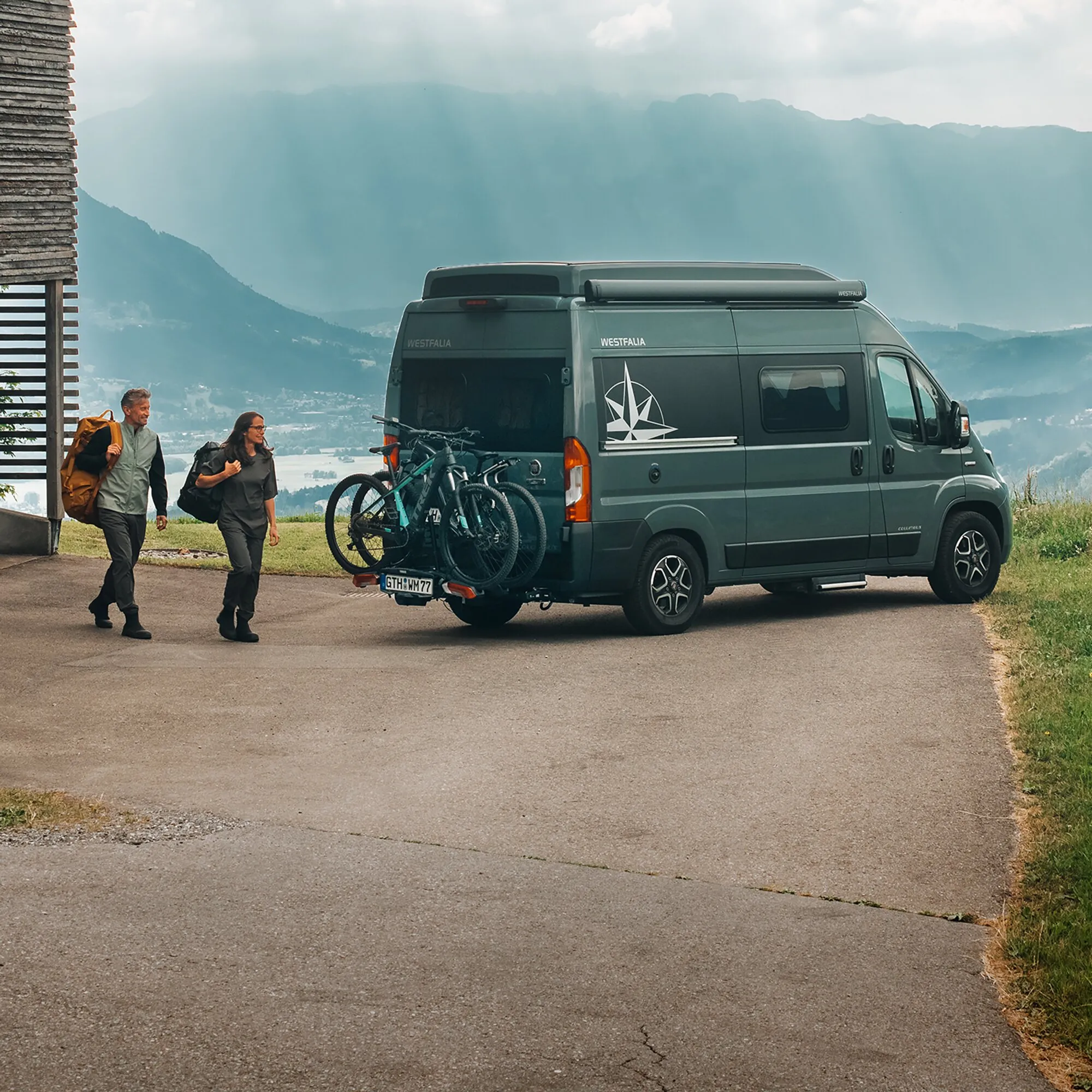 Dunkler Westfalia-Campingvan mit Thule VeloSwing und drei Fahrrädern vor einer alpinen Berglandschaft, zwei Wandernde daneben