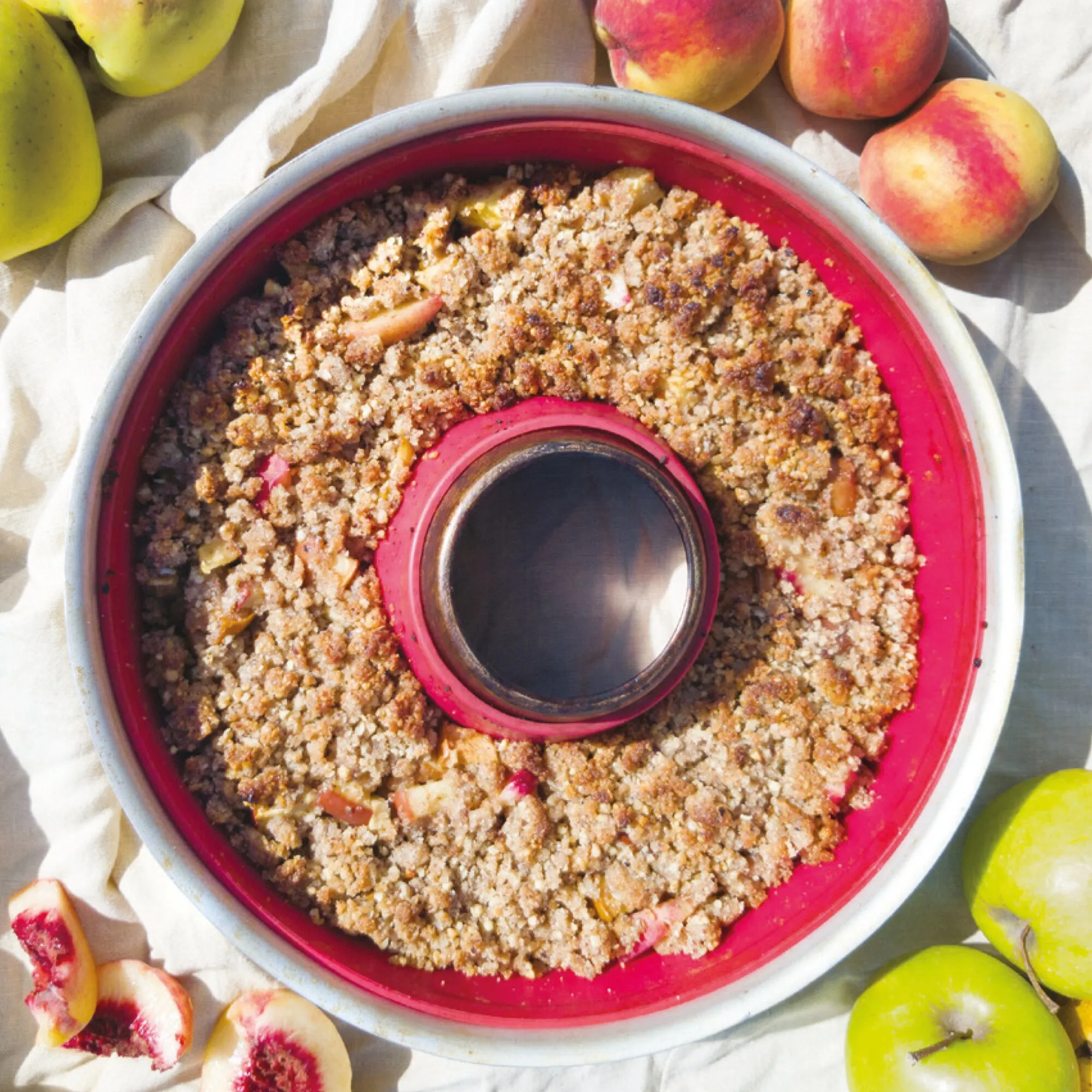 Ringförmiger Streuselkuchen mit Apfel- und Pfirsichstücken im roten Omnia-Backofen, umgeben von frischem Obst aus dem Kochbuch Leicht & Fit Rezepte