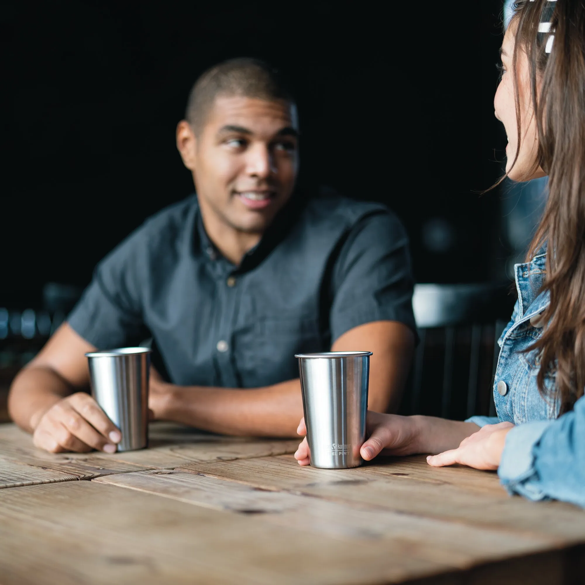Zwei Personen sitzen an einem Holztisch mit silbernen Pint Cups aus Edelstahl, einer davon wird von einer Hand gehalten