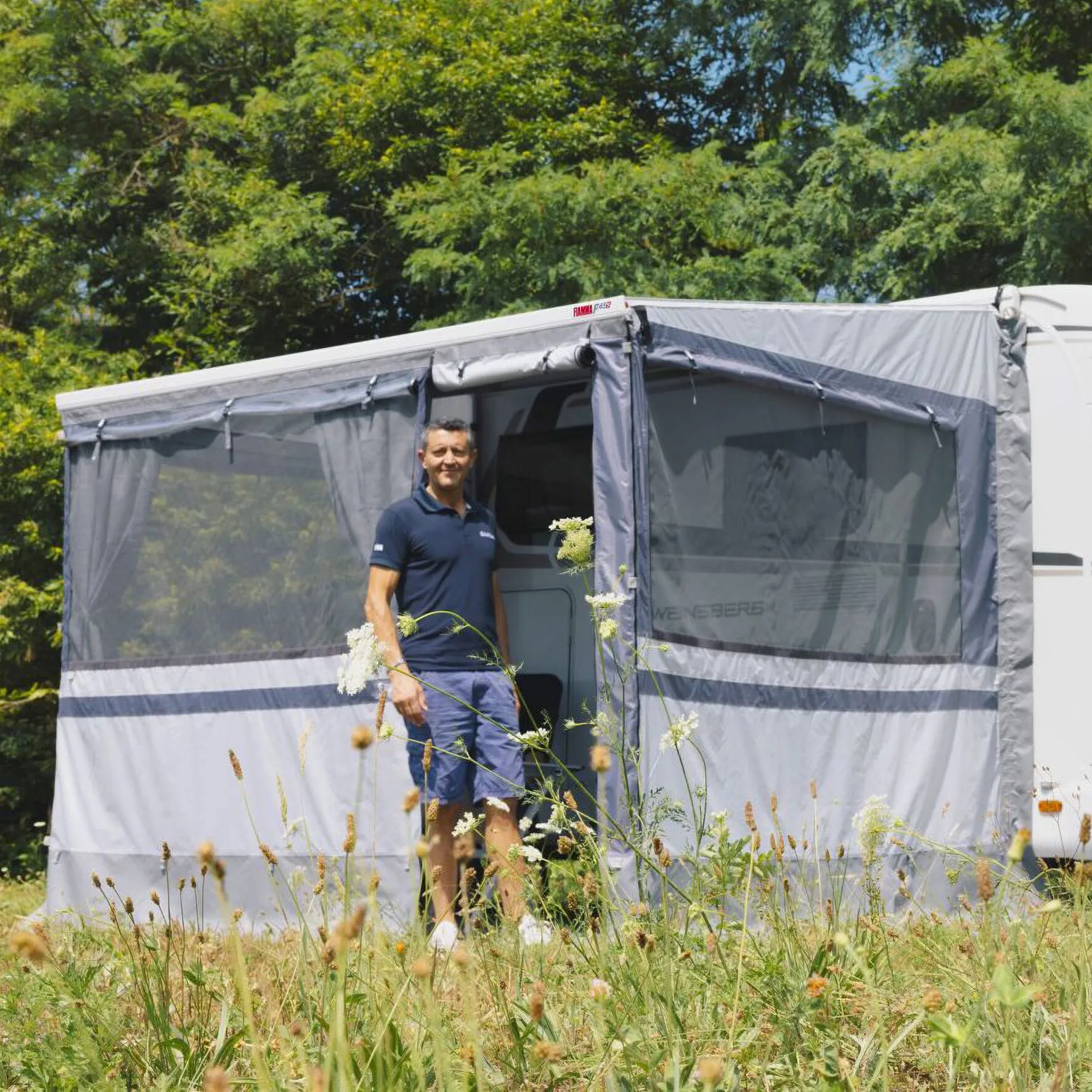Graues Privacy Room CS Light XL, 310 cm, an einer Wohnmobilmarkise mit transparenten Seitenfenstern und geöffneter Tür in einer Wiese