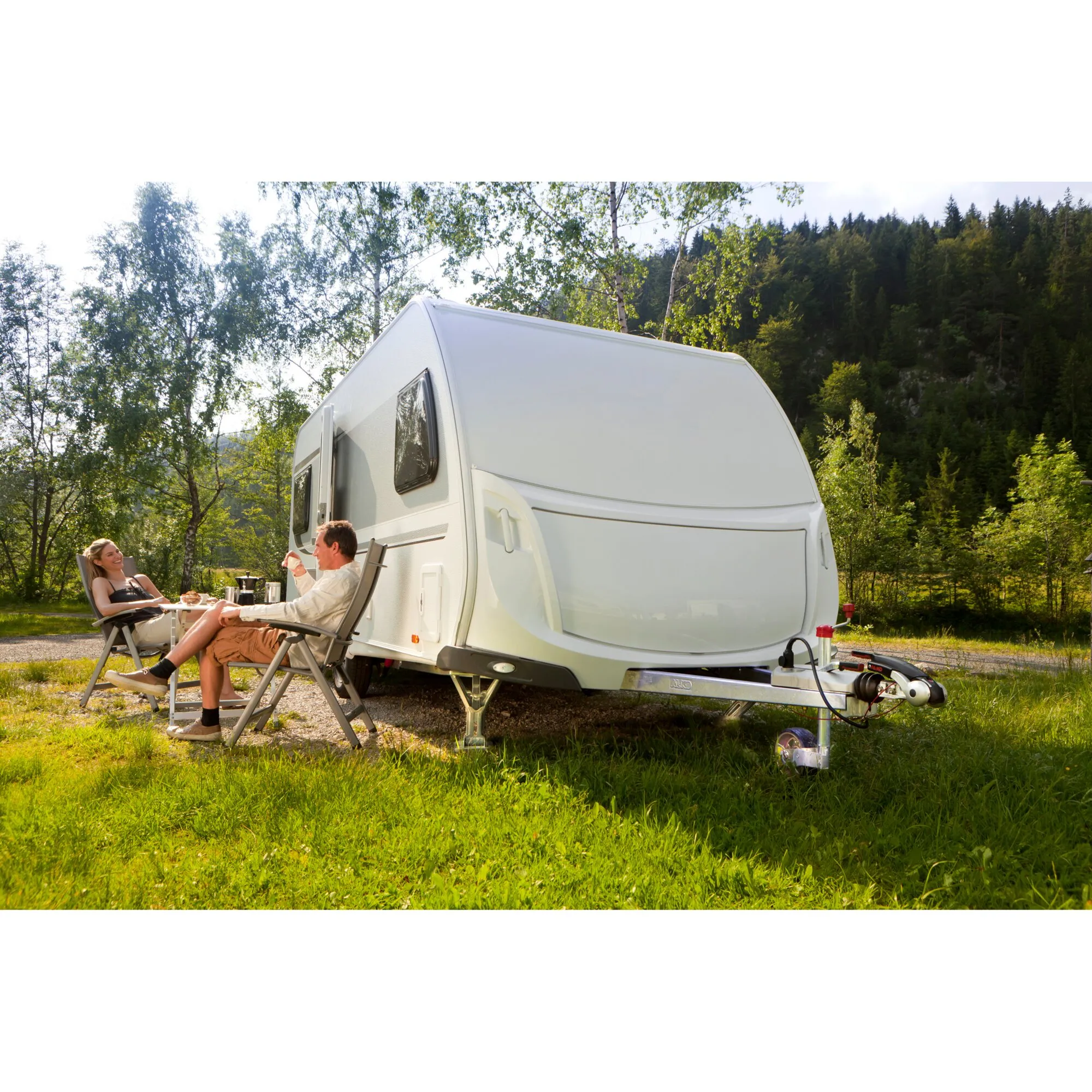 Weißer Wohnwagen mit Sicherheitskupplung AKS 3004 an der Deichsel, zwei Menschen sitzen davor in Campingstühlen im Waldыхәтә