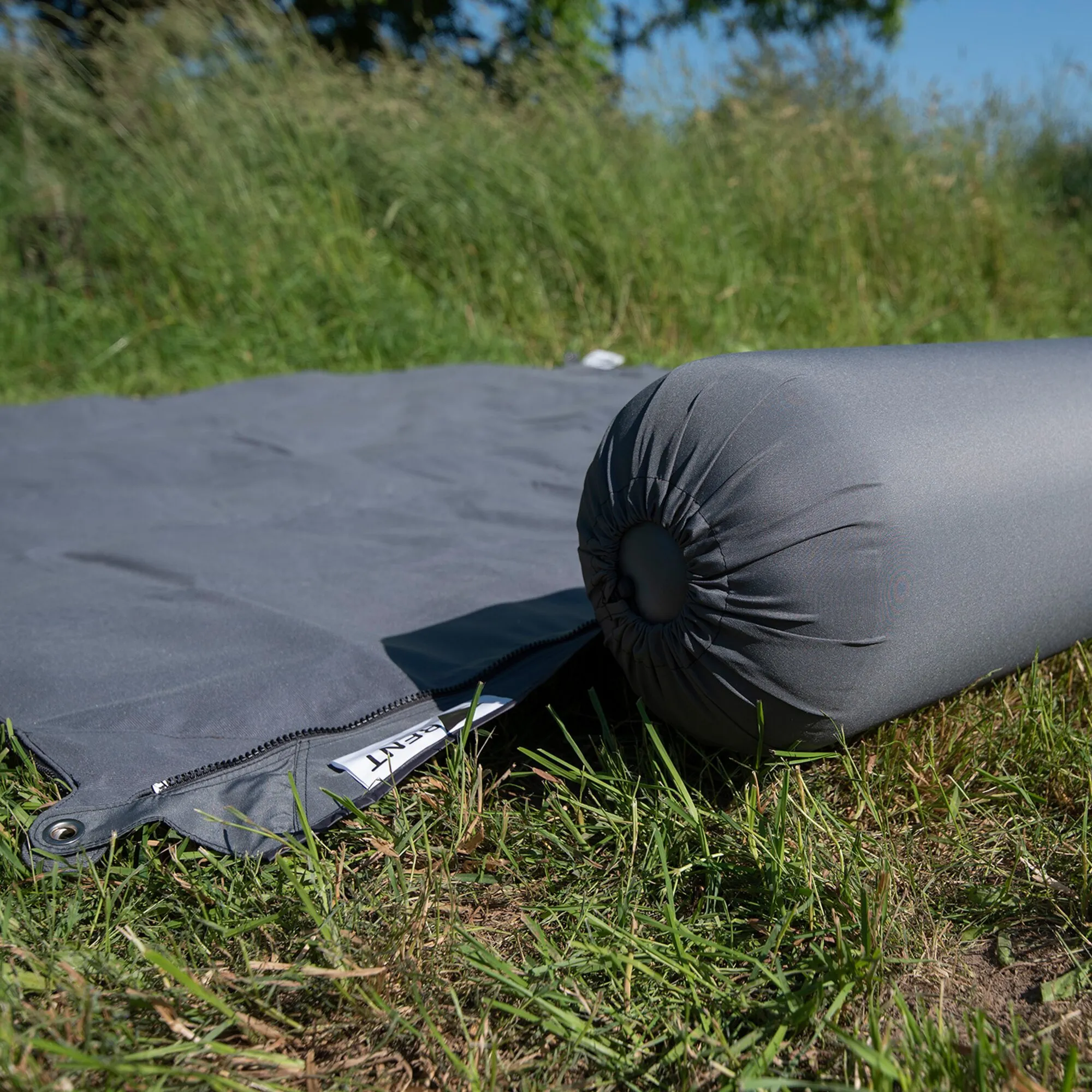 Dunkelgraue Kissenrolle Zip XL Lounger liegt auf einer Wiese; der aufgeklappte Teil mit Reißverschluss ist sichtbar