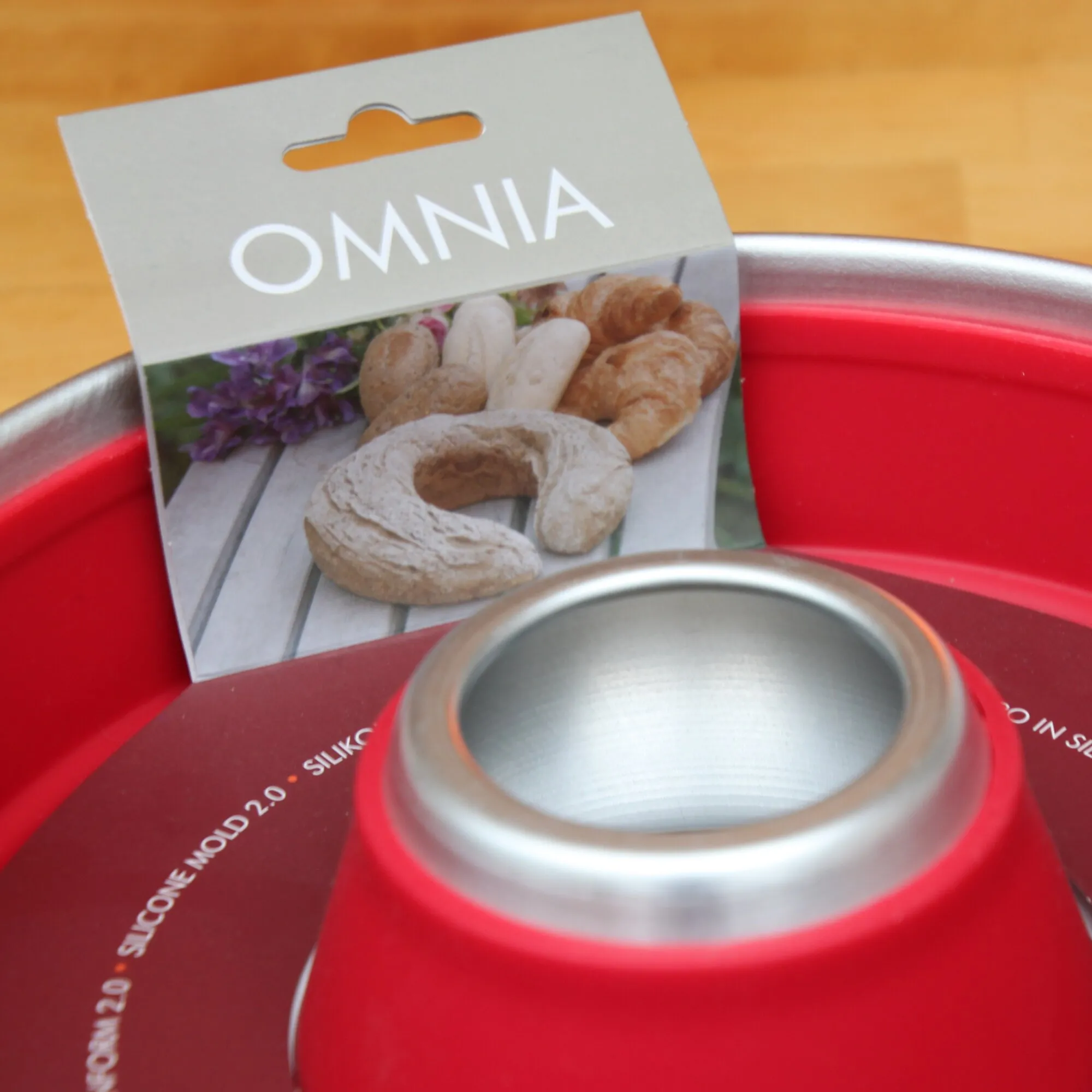 Rote Muffin-Backform aus Metall mit runder Vertiefung und silbernem Rand, dahinter Verpackung mit dem Aufdruck OMNIA