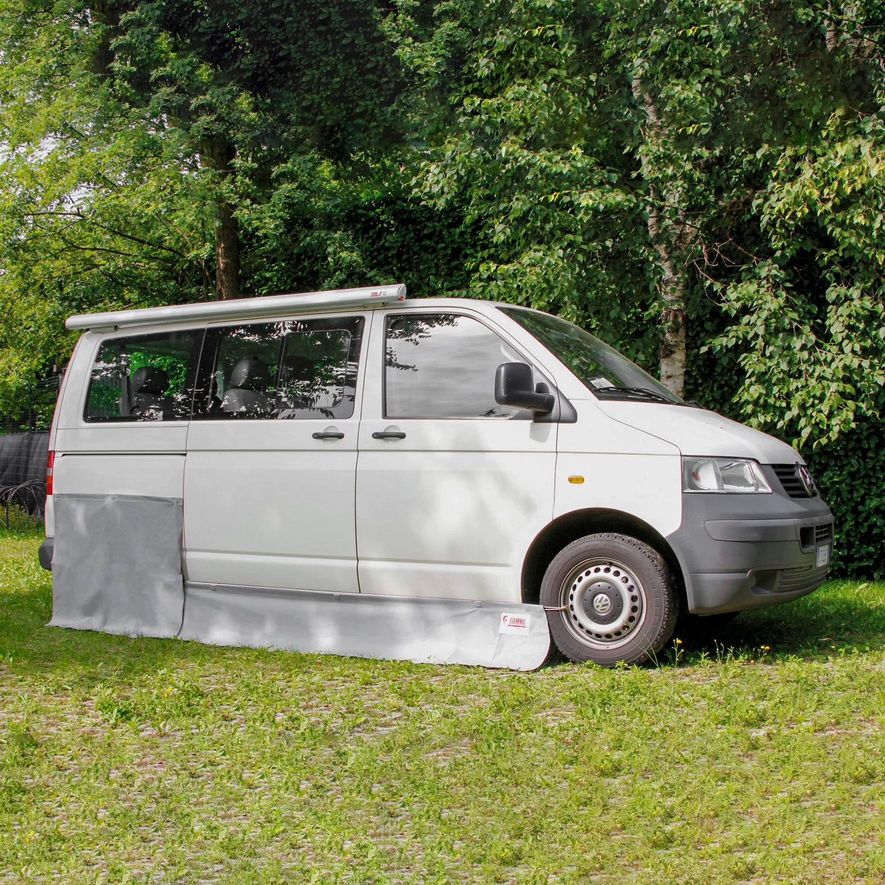Grauer Bodenschutz als Schürze an der Seite eines weißen VW T5/T6 mit Markise im Grünen