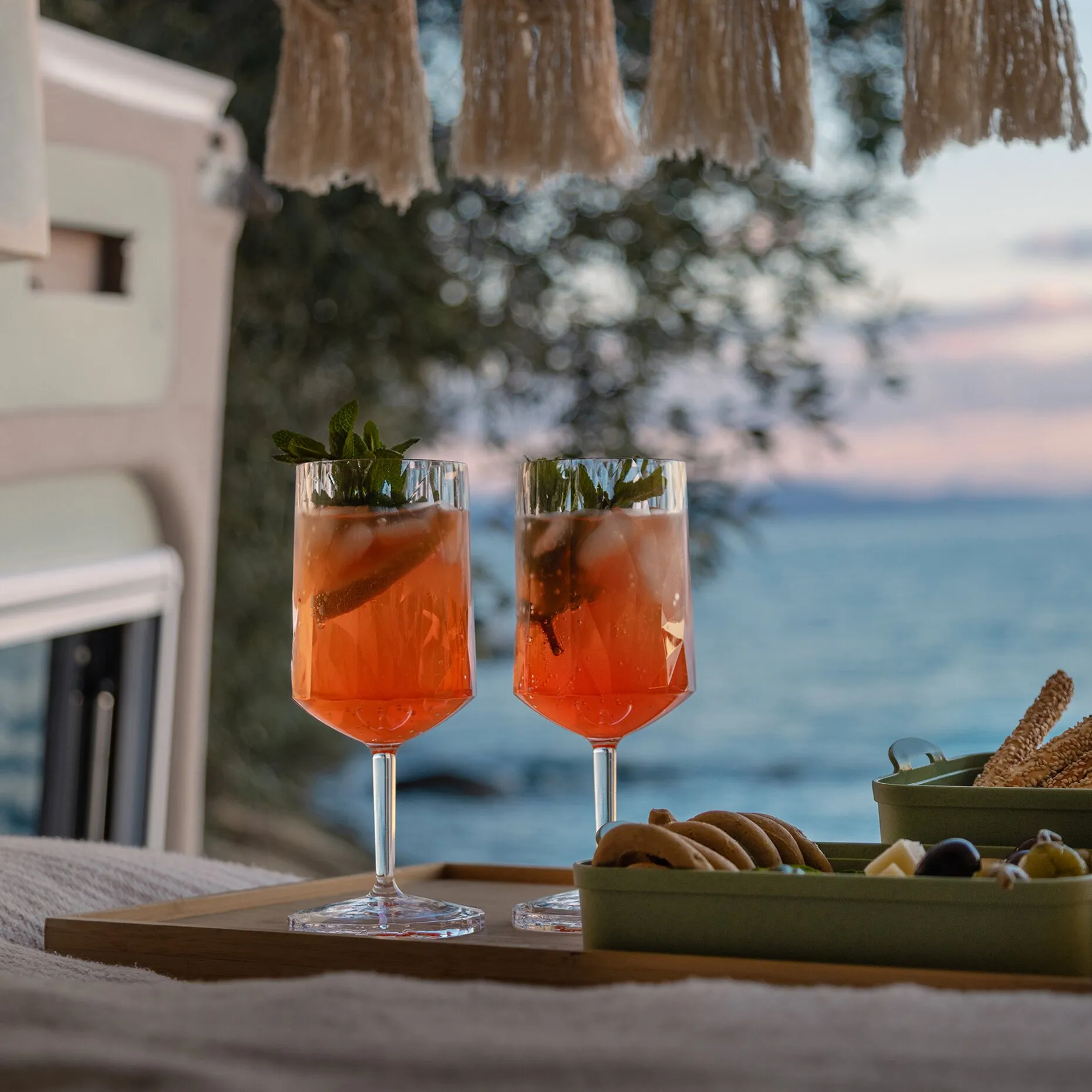 Zwei facettierte Rotweingläser mit orange-rotem Getränk und Minzblättern, auf einem Holztisch am Strand; im Hintergrund Meer und beige Quasten