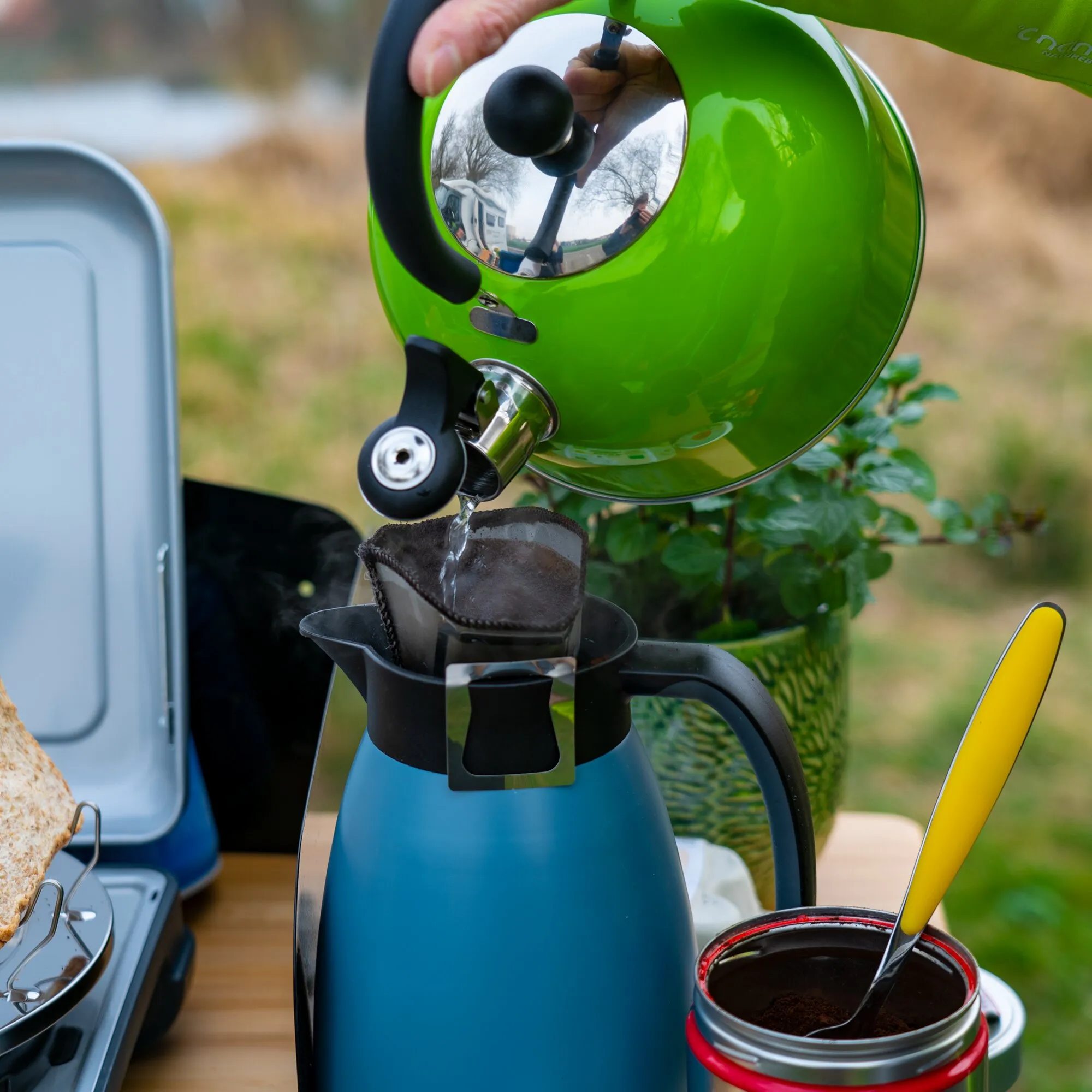 Grüner Kaffeefilter Amigo gießt heißes Wasser in einen Papierfilter über einer blauen Kaffeekanne im Freien