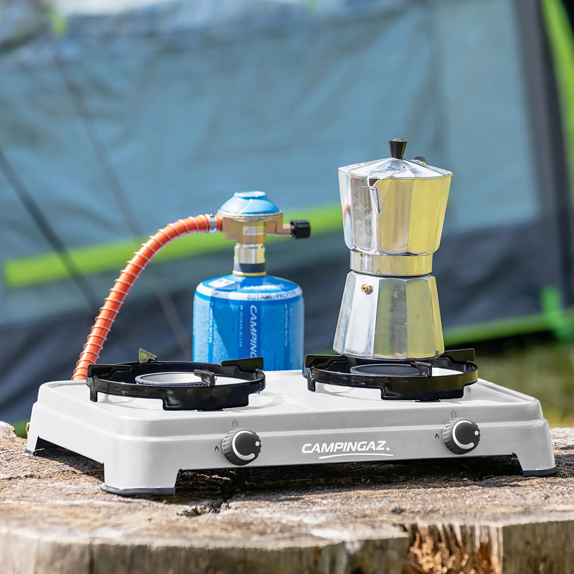 Campingaz Camping Cook CV mit zwei Brennern, blauer Gasflasche und silberner Espressokanne auf einem Campingtisch im Freien