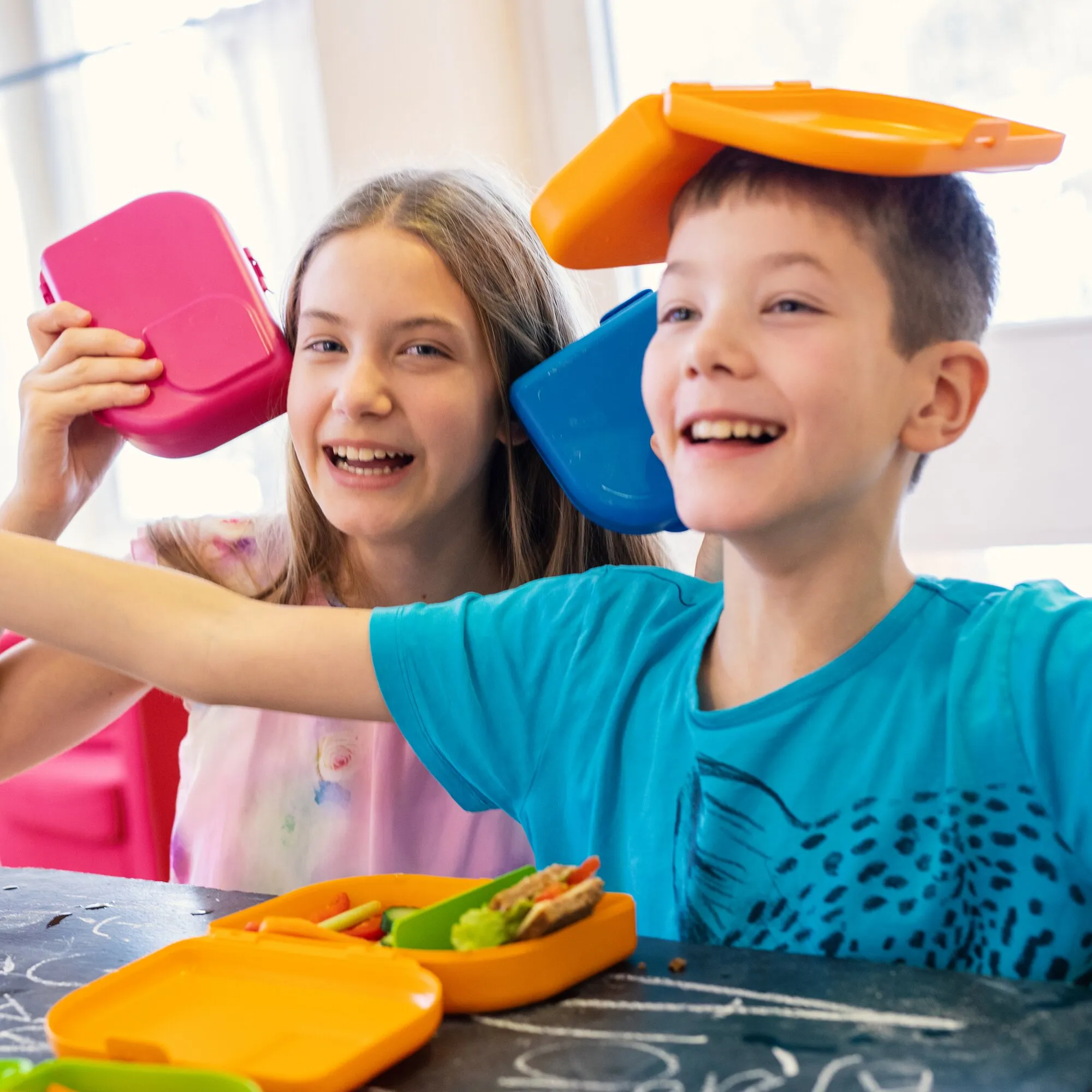 Zwei lachende Kinder mit bunten Brotdosen; der Junge hält eine blaue Brotdose neben sein Gesicht, davor steht eine orange mit Pausenbrot gefüllte Box