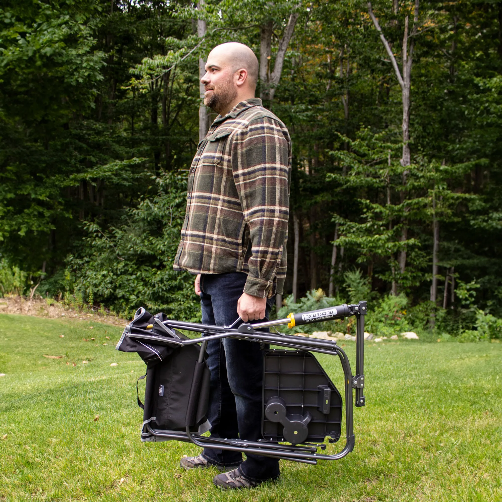 Mann trägt den zusammengeklappten schwarzen Camping-Schaukelstuhl Freestyle Rocker XL™ mit Tisch über eine Rasenfläche im Wald