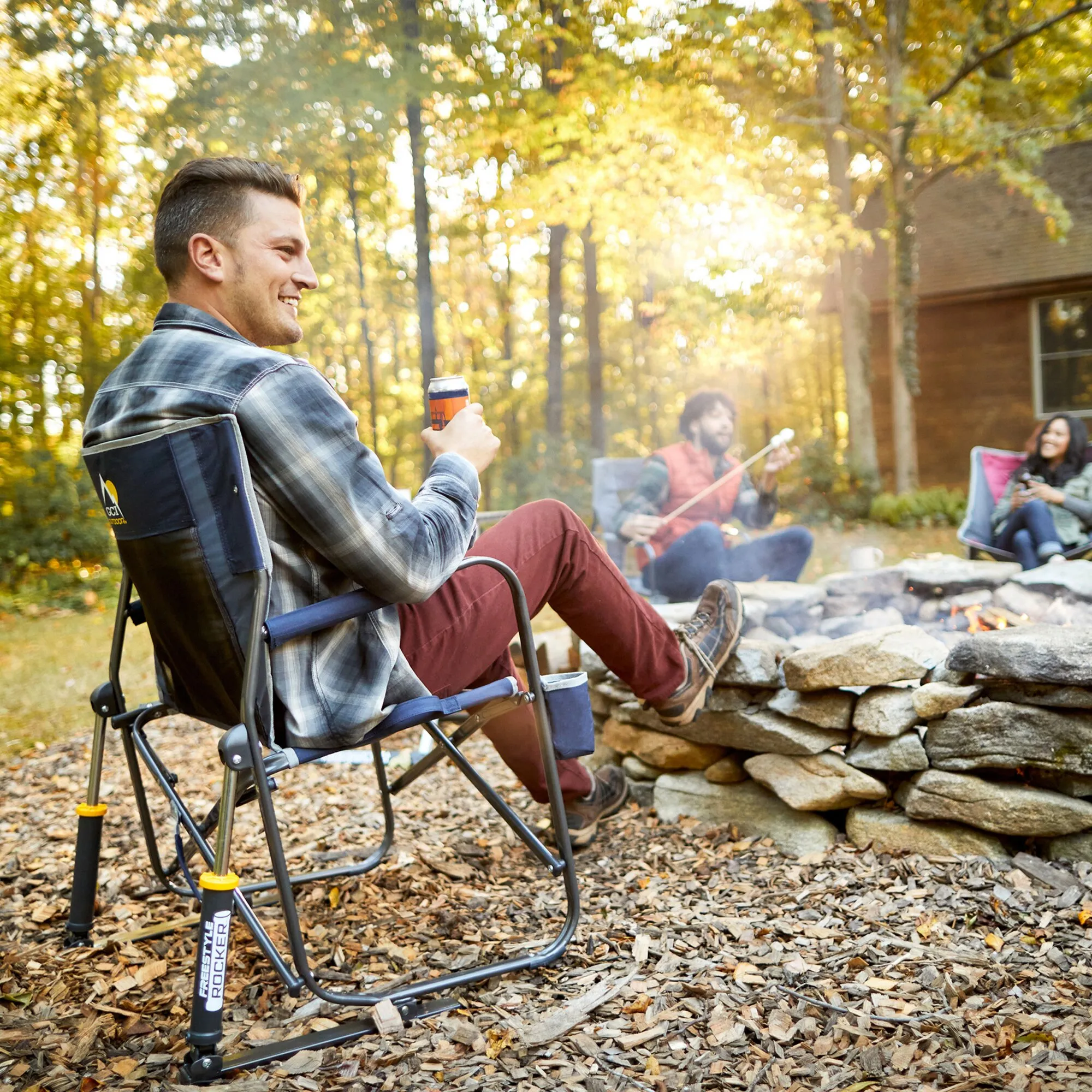 Blauer Camping-Schaukelstuhl Freestyle Rocker mit sitzendem Mann am Lagerfeuer im Wald, Freunde musizieren im Hintergrund