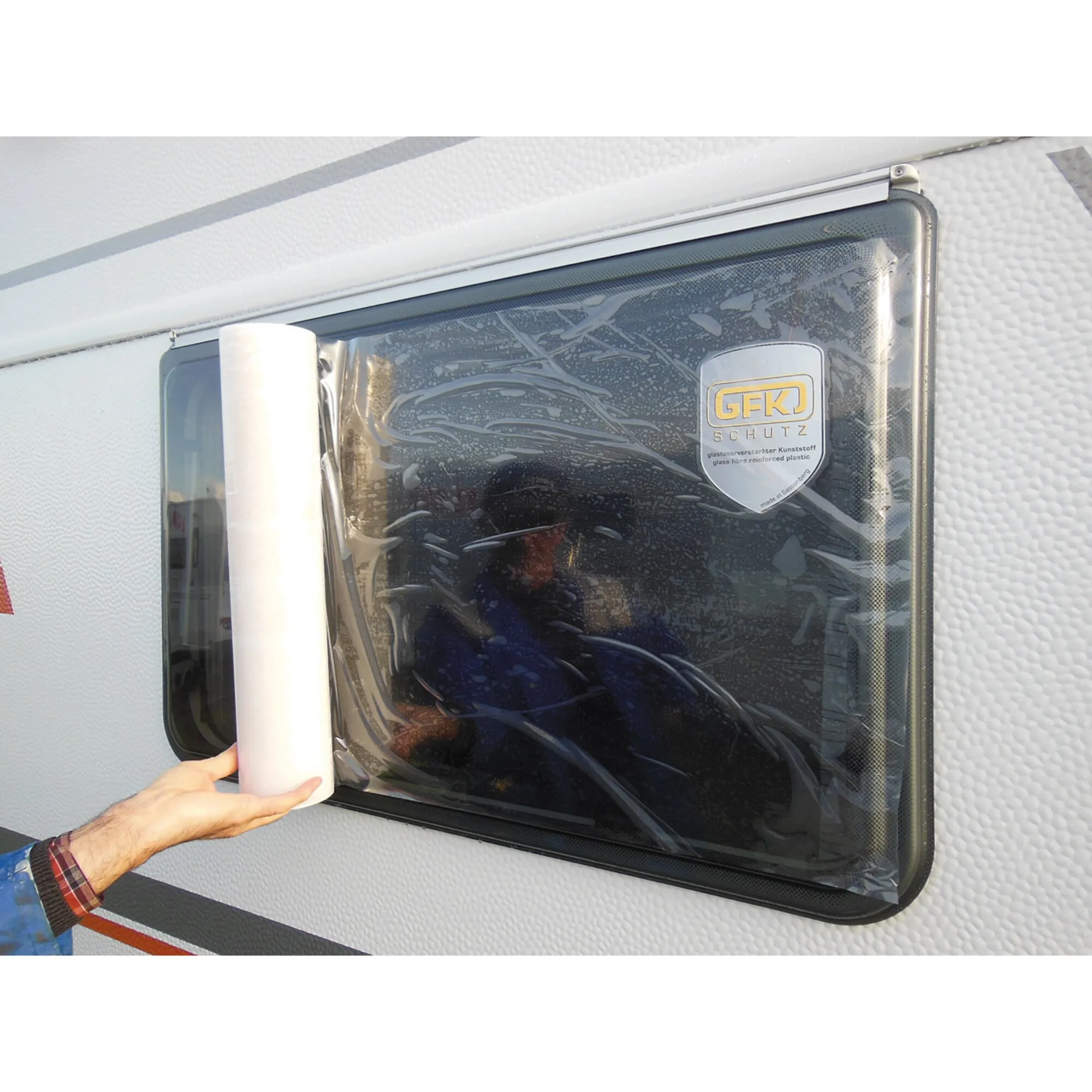 Getönte Fensterschutzfolie wird mit einer Rolle auf ein rechteckiges Fenster aufgebracht; sichtbares GFK-Schutzschild am Fensterrahmen