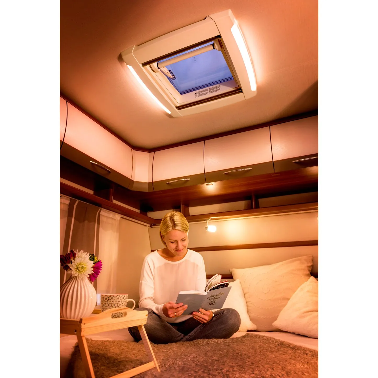 Cremeweiße Seitenblenden VisionStar M mit integrierten LED-Leuchten am Dachfenster im Wohnmobil, darunter liest eine Frau