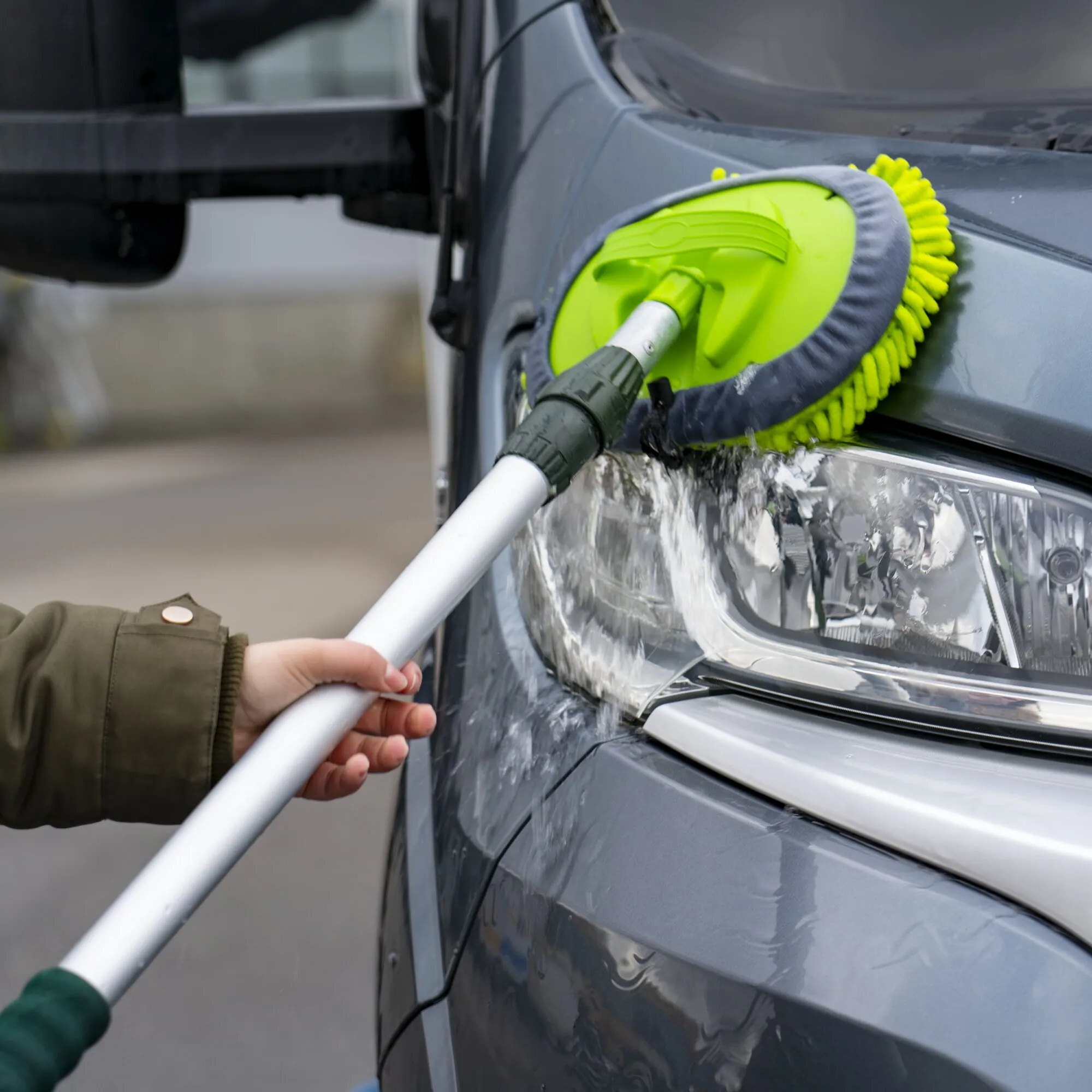 Grüner Mopp-Aufsatz mit grauen Mikrofaserfransen reinigt unter laufendem Wasser die Front eines grauen Autos