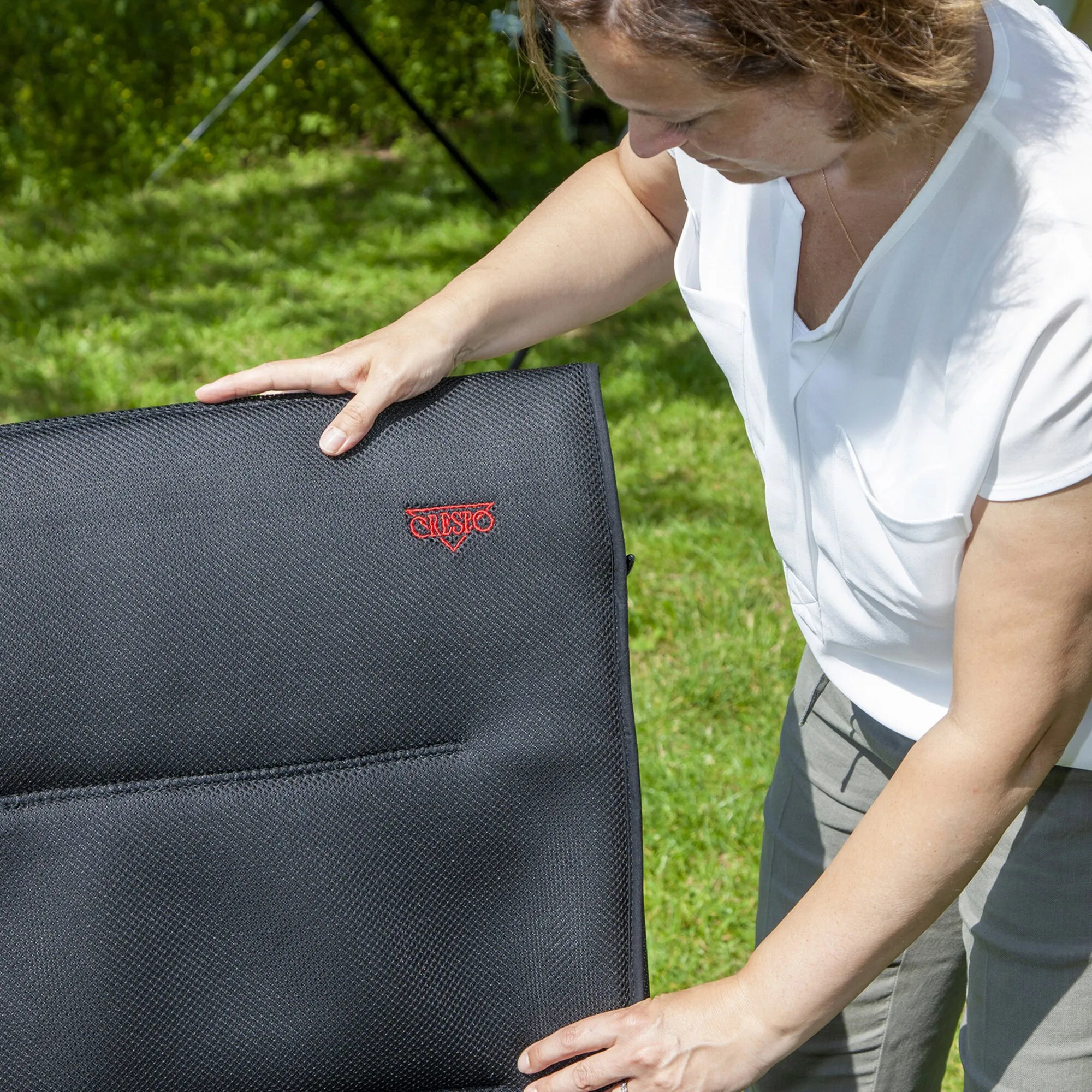 Frau positioniert eine schwarze Polsterauflage Air-Deluxe mit rotem Crespo-Logo im Garten grasbewachsenen Außenbereich